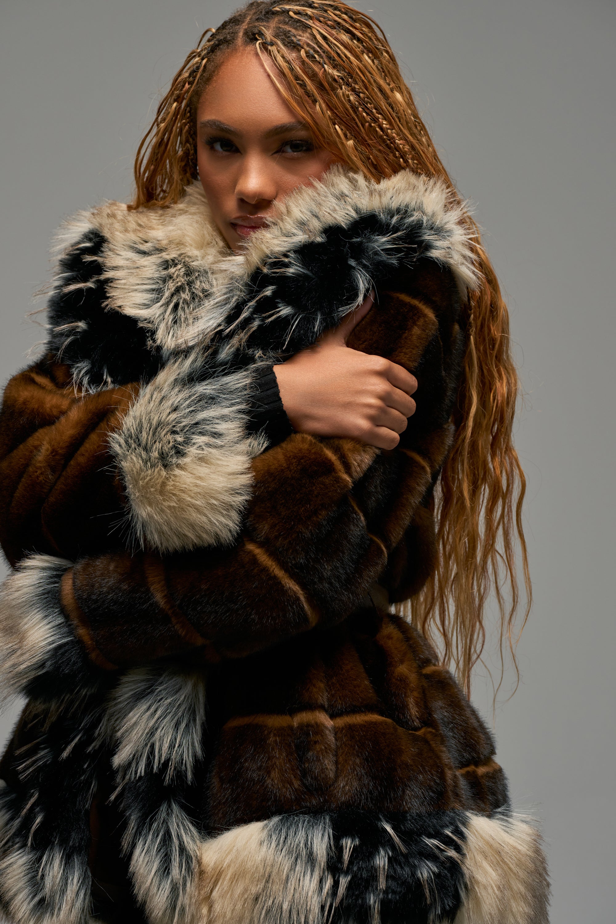 Wearing the KENZ FAUX FUR JACKET, which features a luxurious mix of black, brown, and white sections, a person with long braided hair poses with arms crossed against a plain gray background.