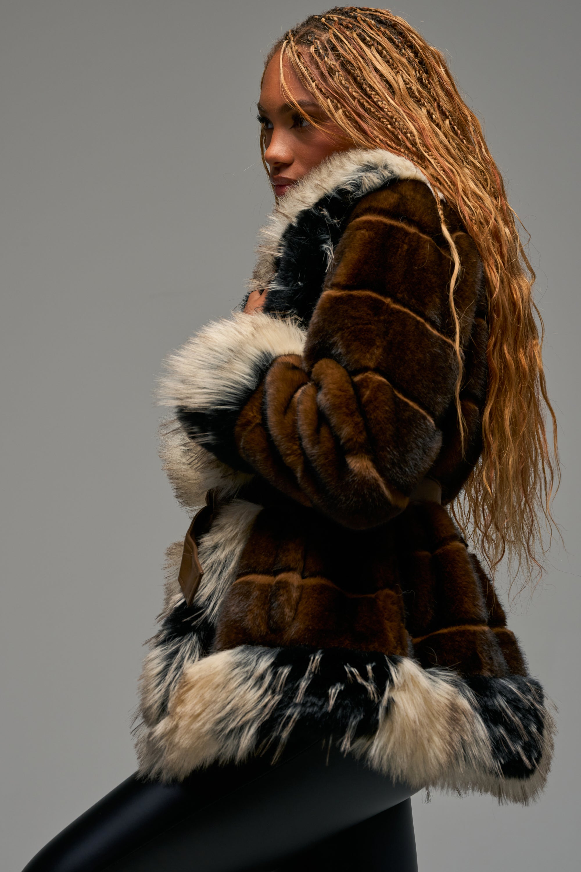 Wearing the KENZ FAUX FUR JACKET and black leggings, a woman with long braided hair poses in profile against a plain gray background, showcasing chic winter styling.