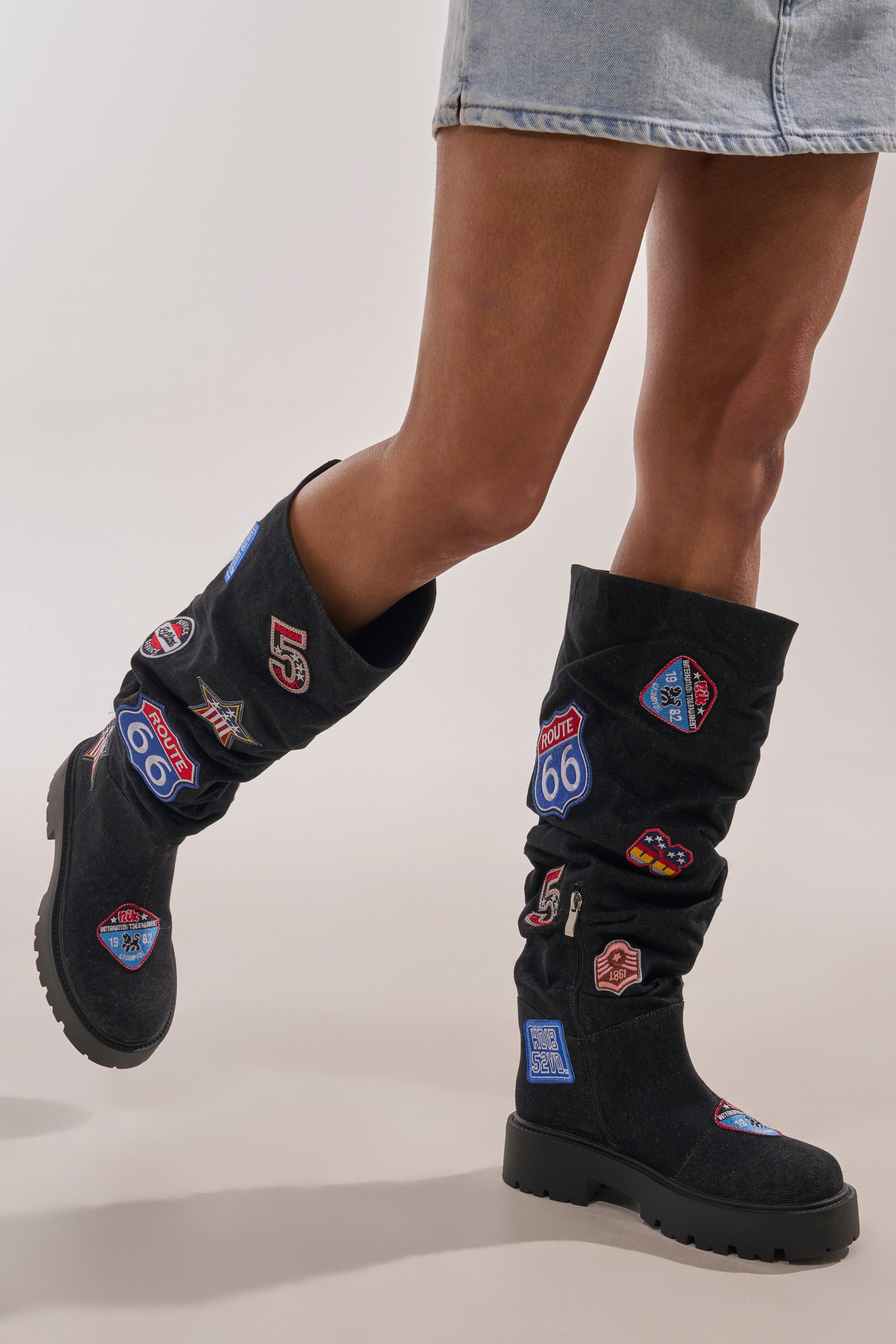 A person wears a light denim skirt and AKIRA GOLD POSTAL BLACK PATCHWORK FLAT BOOT, featuring colorful route and highway patches, standing with one leg slightly bent against a plain light-colored background. 0