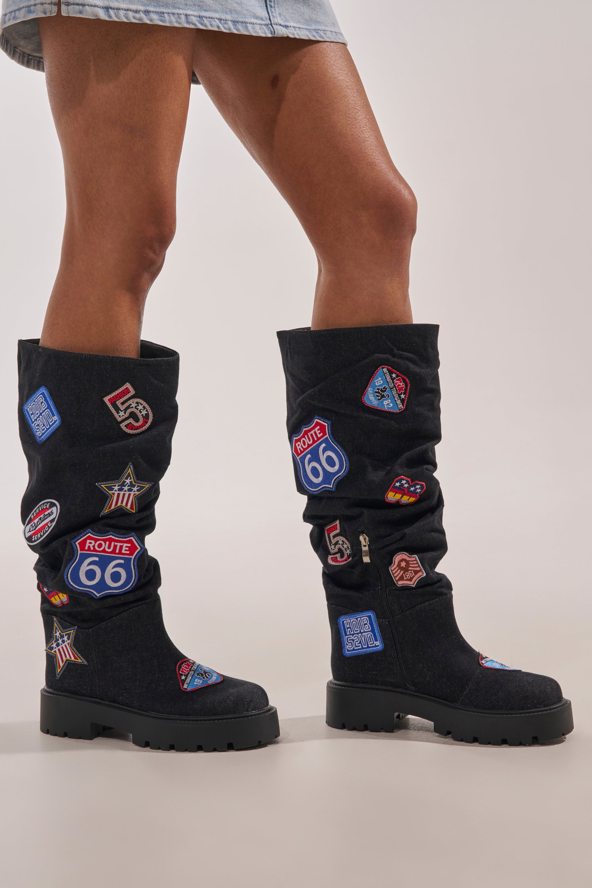 A person stands against a plain background wearing a light denim skirt and the AKIRA GOLD POSTAL BLACK PATCHWORK FLAT BOOT. 1