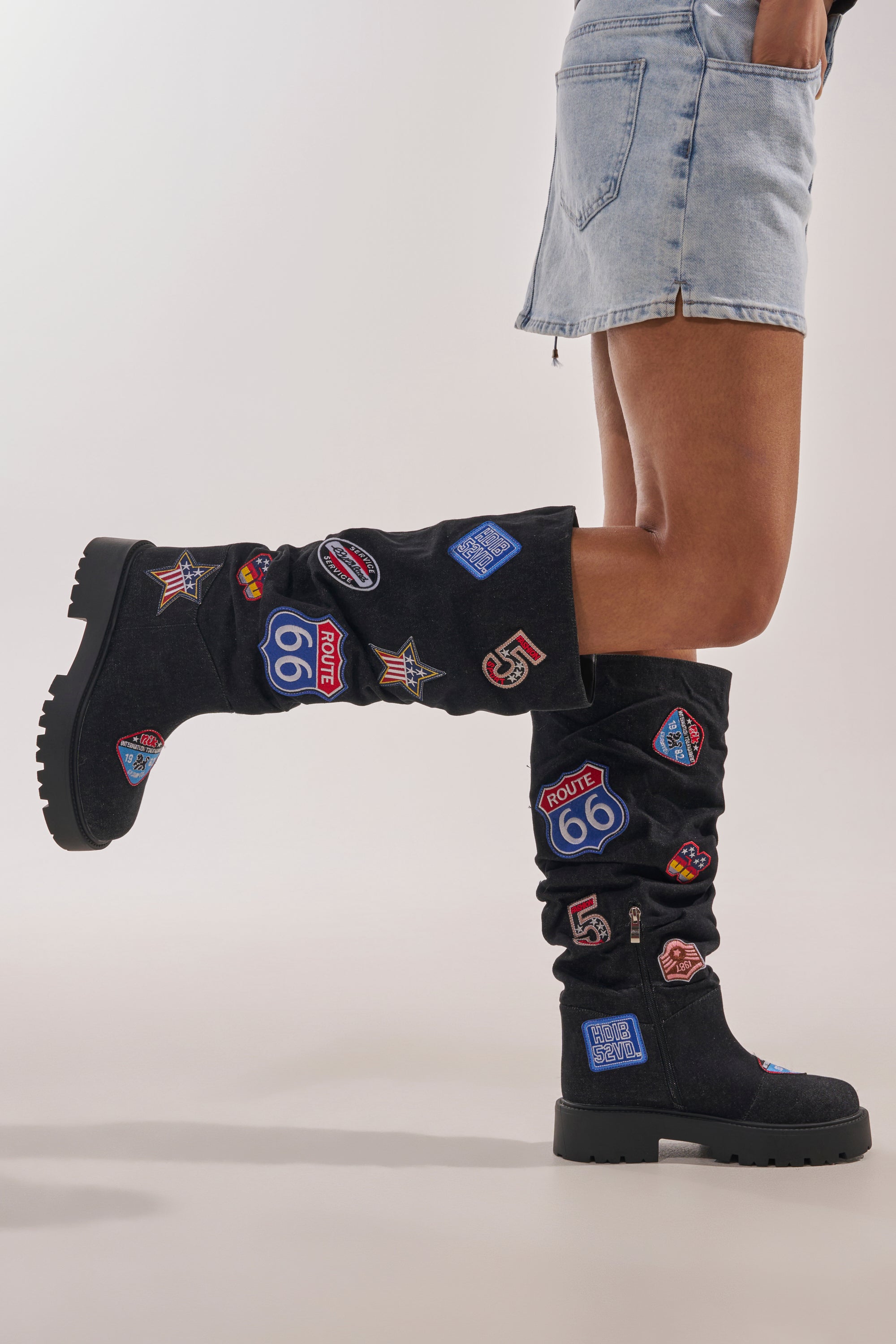 A person wearing a light denim mini skirt and AKIRA GOLD POSTAL BLACK PATCHWORK FLAT BOOT with colorful Route 66 patches stands with one knee bent, highlighting the boots' unique embroidered designs. 2