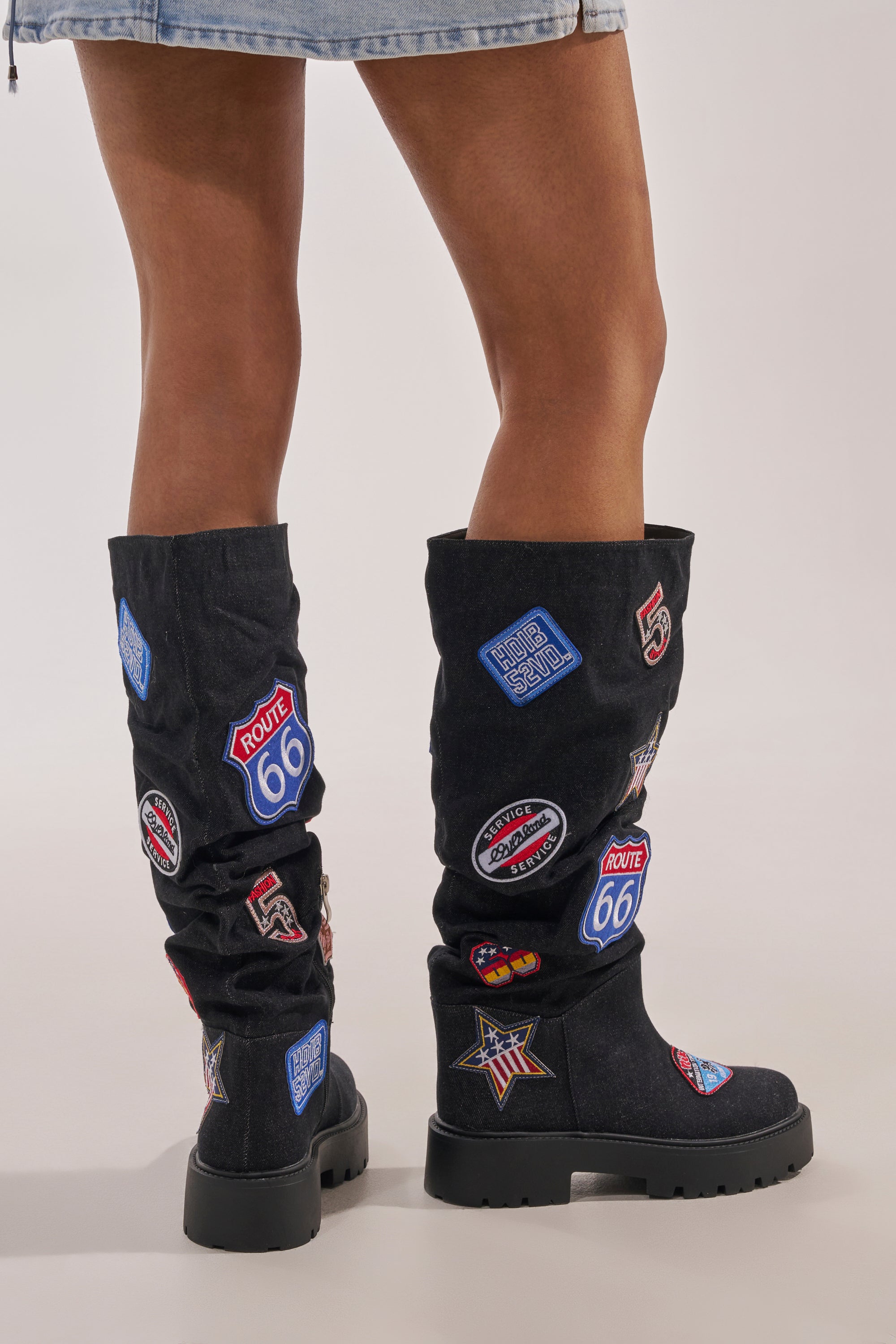A person stands on a light background wearing the AKIRA GOLD POSTAL BLACK PATCHWORK FLAT BOOT, slouchy knee-high black boots decorated with colorful patches like Route 66, paired with a frayed denim skirt. 3