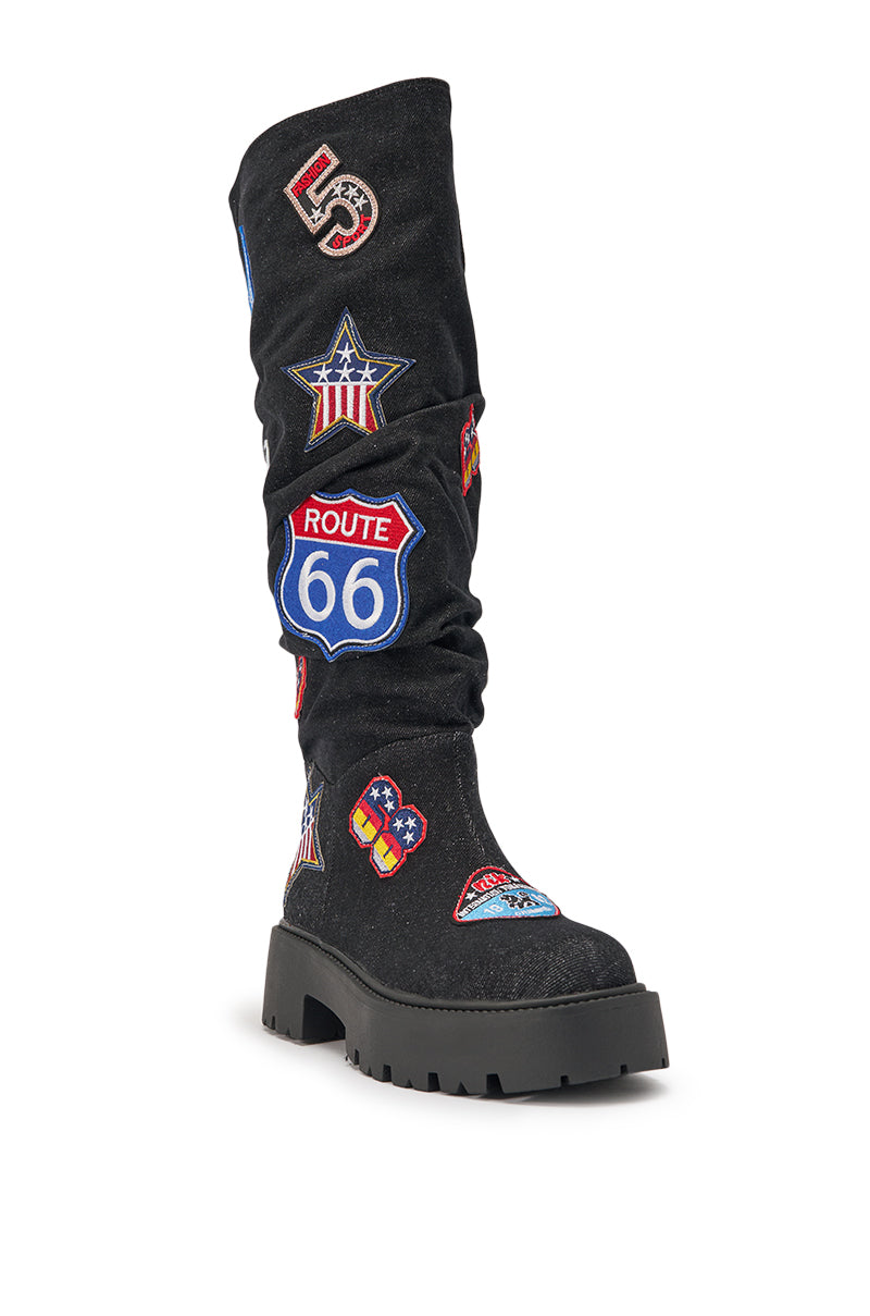 The AKIRA GOLD POSTAL BLACK PATCHWORK FLAT BOOT is a black knee-high boot decorated with colorful postal-themed embroidered patches, including Route 66 signs, stars, and badges, for a bold statement look. 6