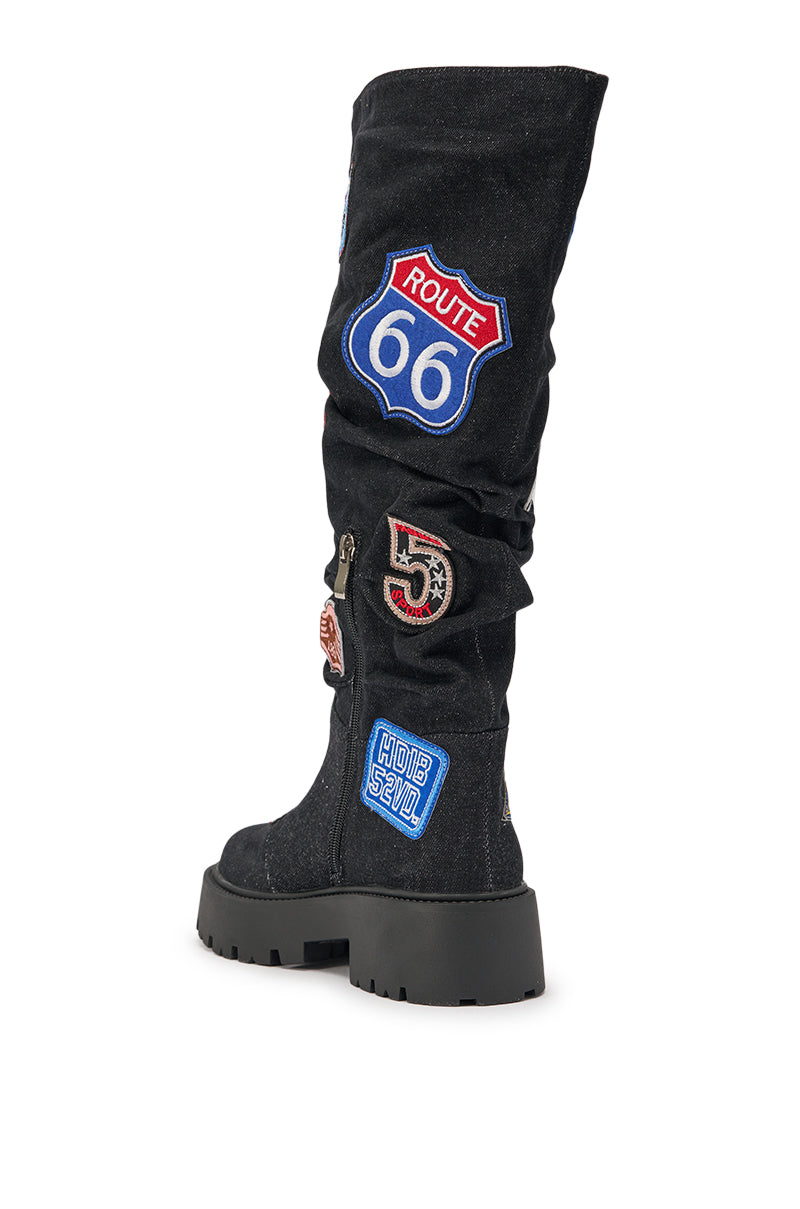 The AKIRA GOLD POSTAL BLACK PATCHWORK FLAT BOOT is a black slouchy knee-high boot with a chunky sole, featuring colorful postal-inspired embroidered patches like "Route 66," the number 5, and a bold "HIDE & SEEK" label. 7