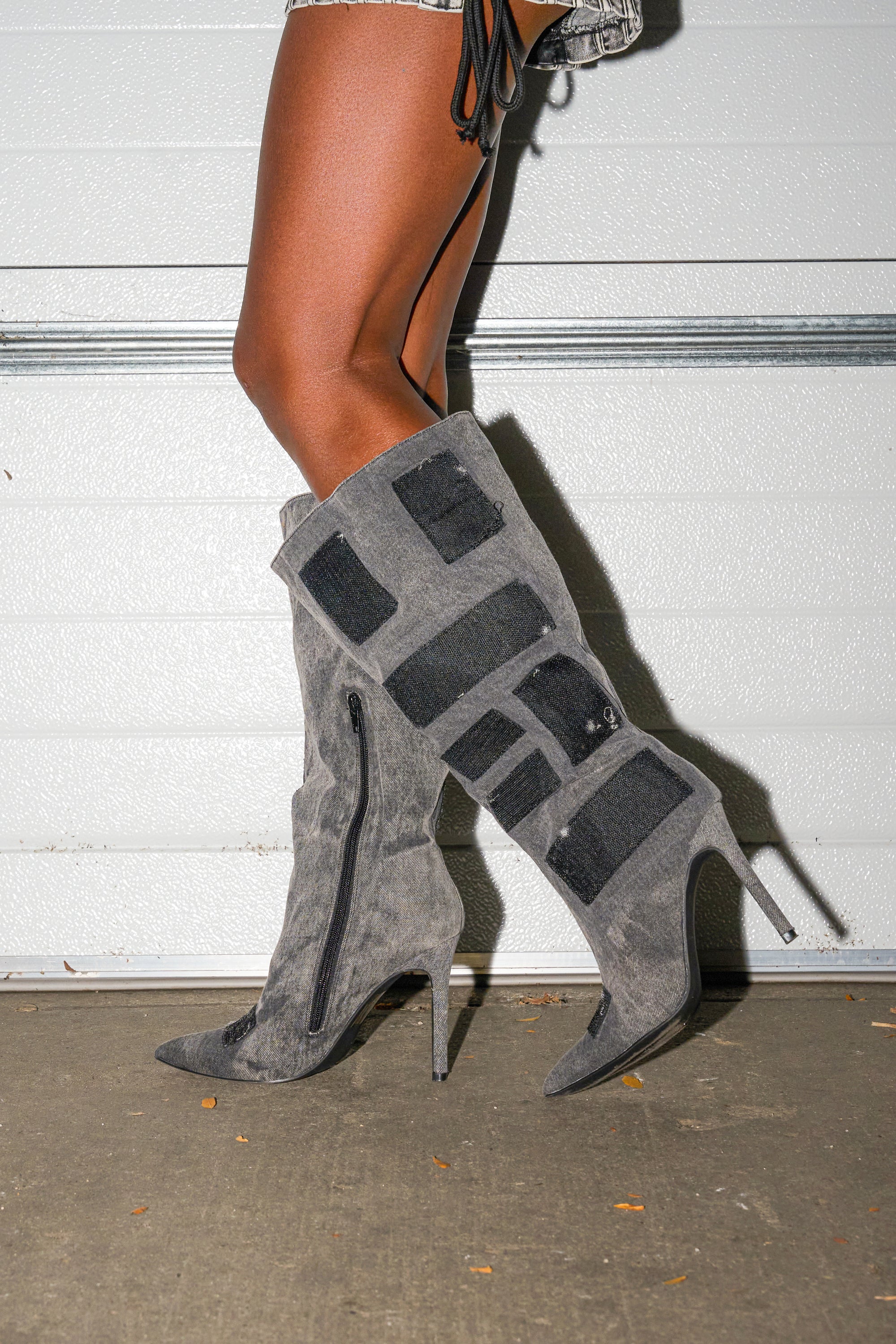 Only the legs are visible as a person stands on concrete in front of a white garage door, wearing AZALEA WANG PRAVIN BLACK POCKET DETAIL STILETTO BOOTS.