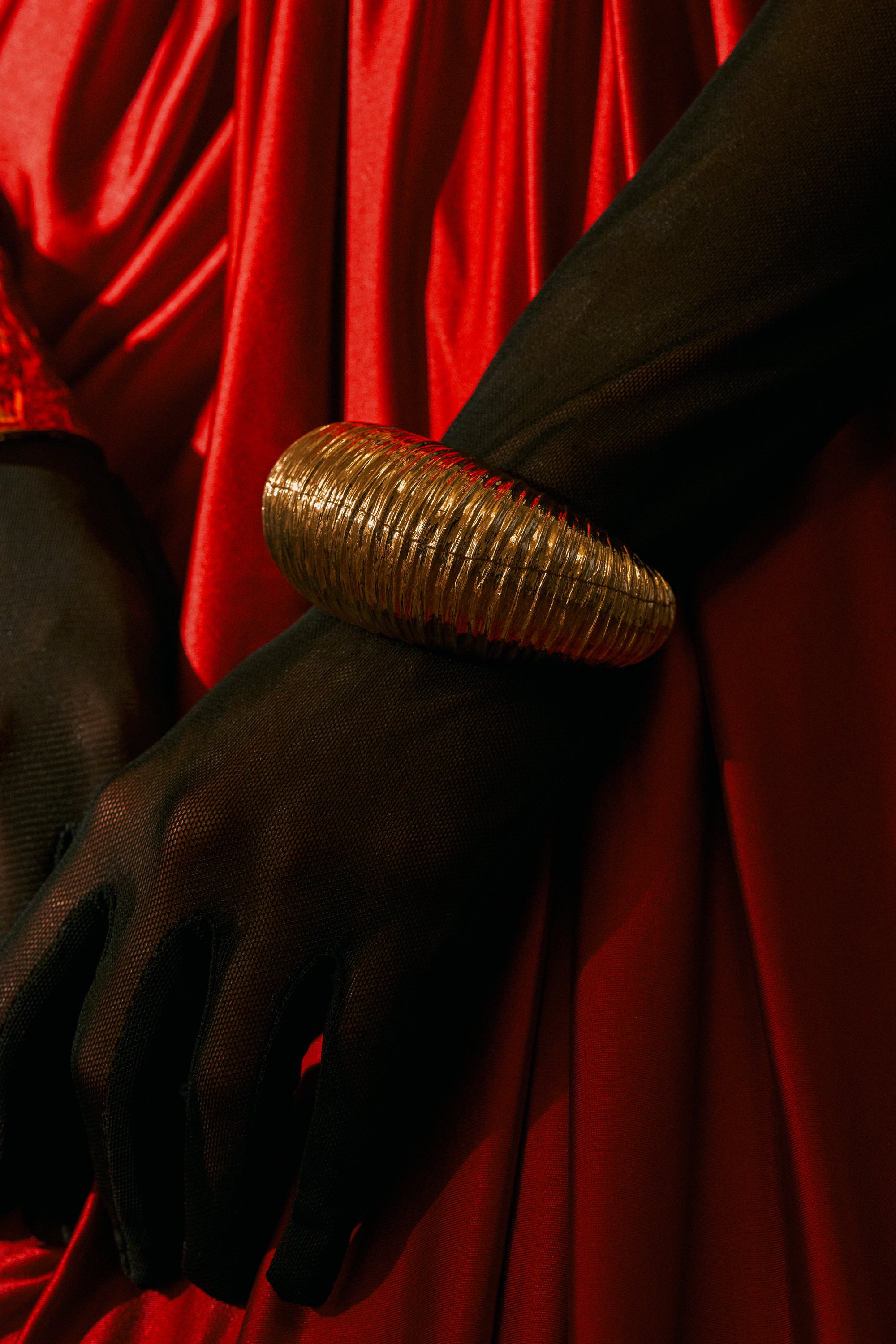 A person wearing black mesh gloves and the LOUDER NOW OVERSIZED BANGLE IN GOLD rests their hand on red satin fabric.