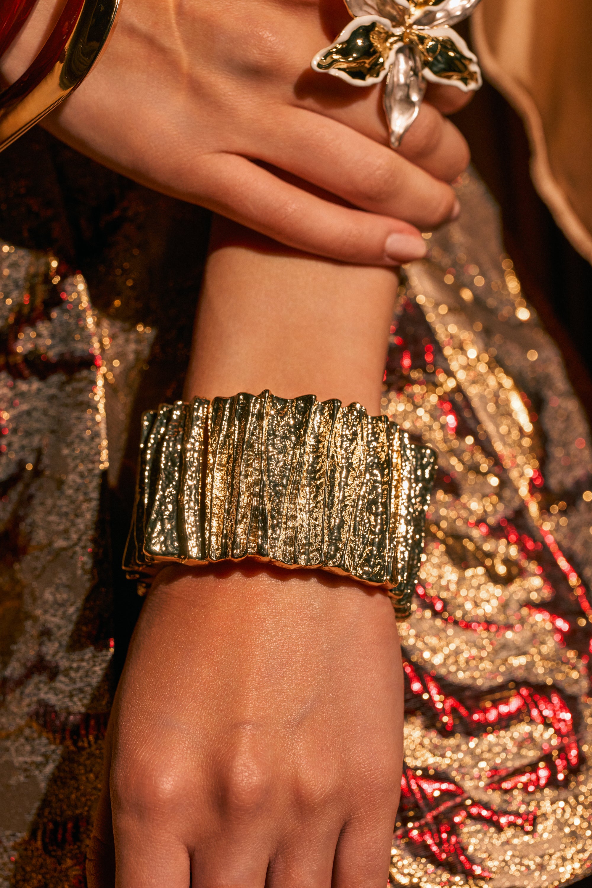 A close-up of a hand wearing the STONE COLD BANGLE IN GOLD, a large sculptural gold cuff bracelet, paired with a bold floral ring against an ornate backdrop of shimmering gold and red fabric.