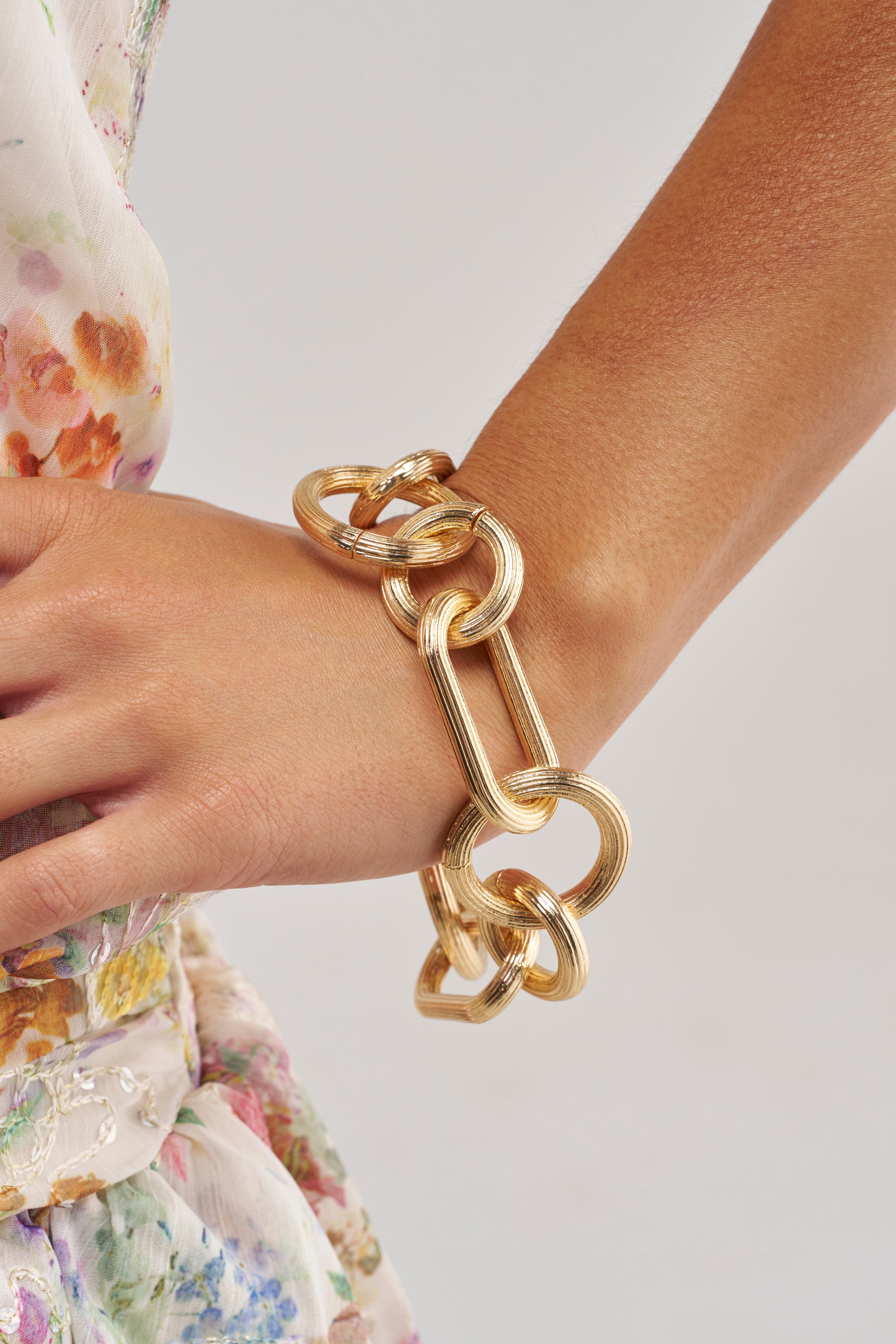 A person wearing the CAN’T BEAT MY OUTFIT CHAIN BRACELET IN GOLD on their wrist, shown against a floral-patterned, light-colored garment—an eye-catching piece of streetwear jewelry that effortlessly blends bold style with elegance.