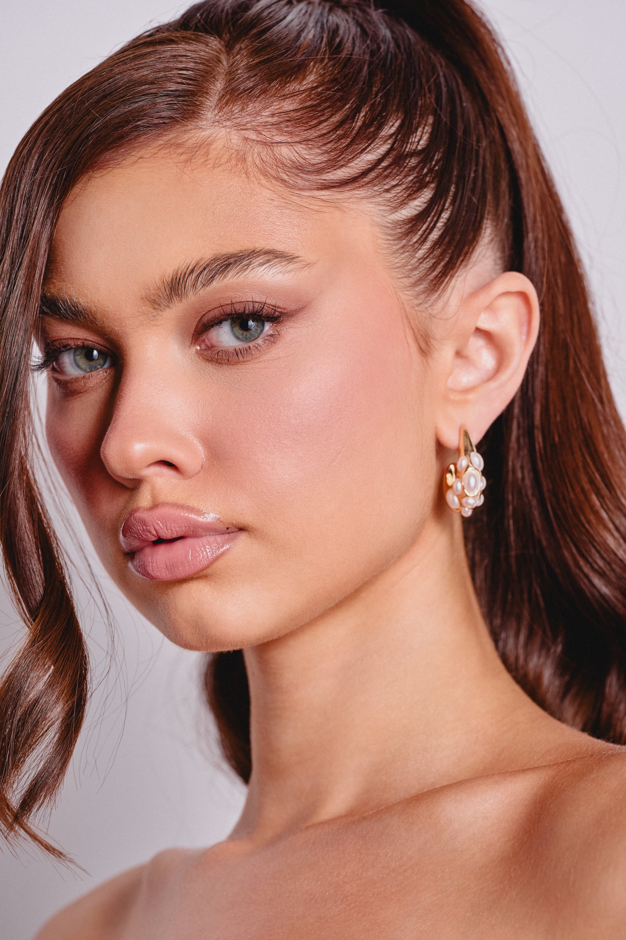 A woman with long brown hair in a high ponytail wears TO THE WATER EARRING, featuring chunky gold and faux pearls. She has defined brows, subtle pink makeup, glossy lips, and faces the camera with a neutral expression. 0