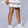 A person wearing a white ruffled skirt and matching PRINCESS WHITE EMBELLISHED SNEAKER with white socks. The image is focused on the legs and feet against a light grey background.