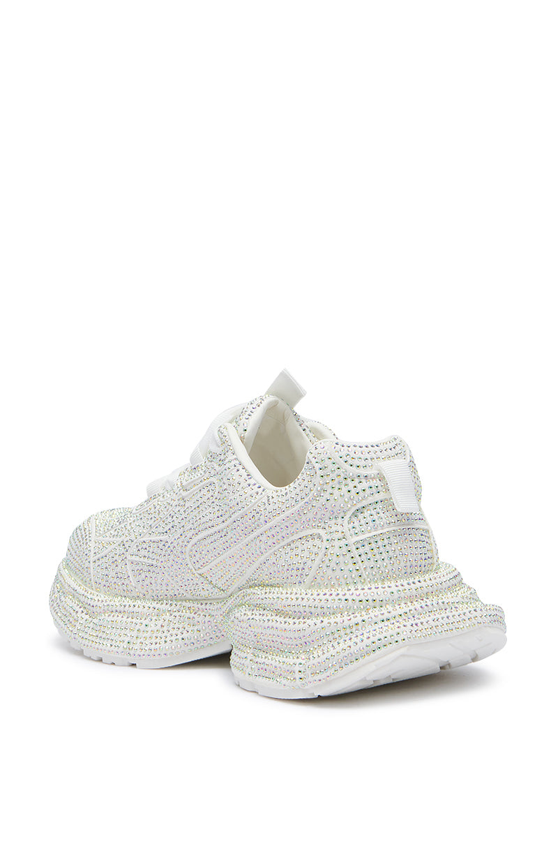 The PRINCESS WHITE EMBELLISHED SNEAKER is a chunky pair of sneakers adorned with sparkling rhinestones. These white shoes feature thick flatform soles and a convenient pull-on tab at the back for easy wearing. With a flashy design intended to catch the light, these rhinestone sneakers are presented against a plain white background.