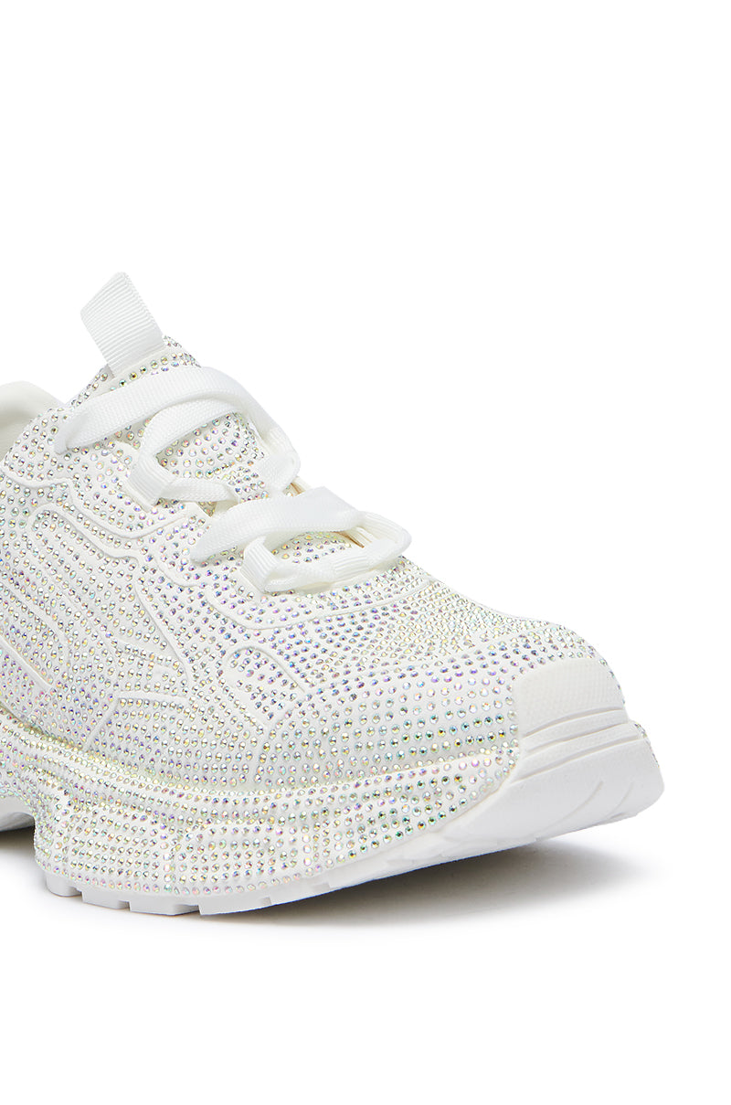 A close-up of the PRINCESS WHITE EMBELLISHED SNEAKER showcases its surface adorned with small, glittering rhinestones. The sneaker features white laces and a thick, textured sole. Its stylish and eye-catching design reflects light to create a sparkling effect.