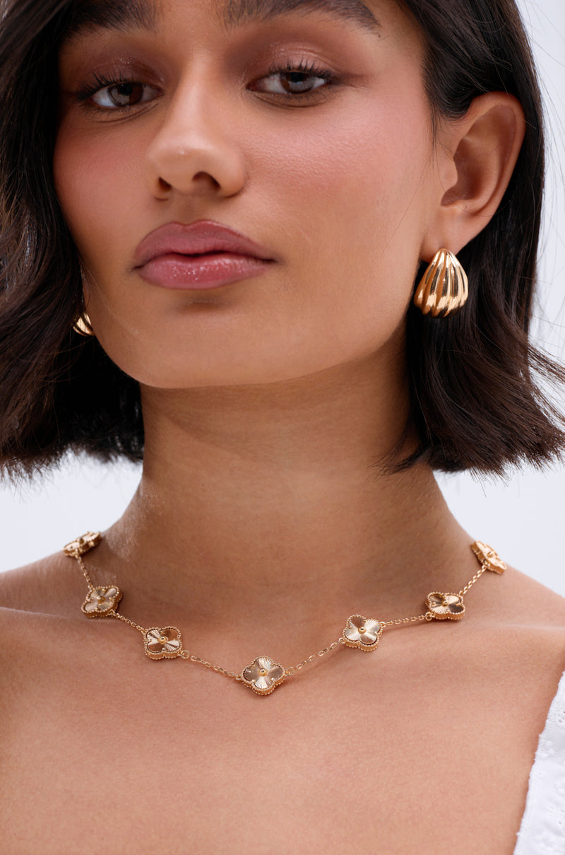 A woman with short dark hair wears gold drop earrings and the QUEZ NECKLACE, a gold-tone piece adorned with floral charms, for a feminine touch. She sports a neutral expression and a white top. 1
