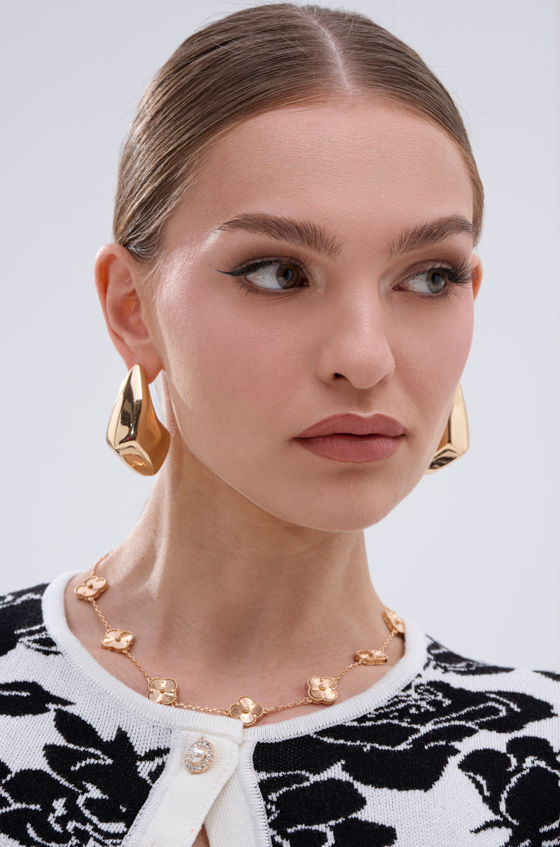A woman with slicked-back hair wears large gold statement earrings and the QUEZ NECKLACE, a gold-tone piece with floral charms. She has natural makeup with winged eyeliner and wears a black-and-white floral-patterned top. 2