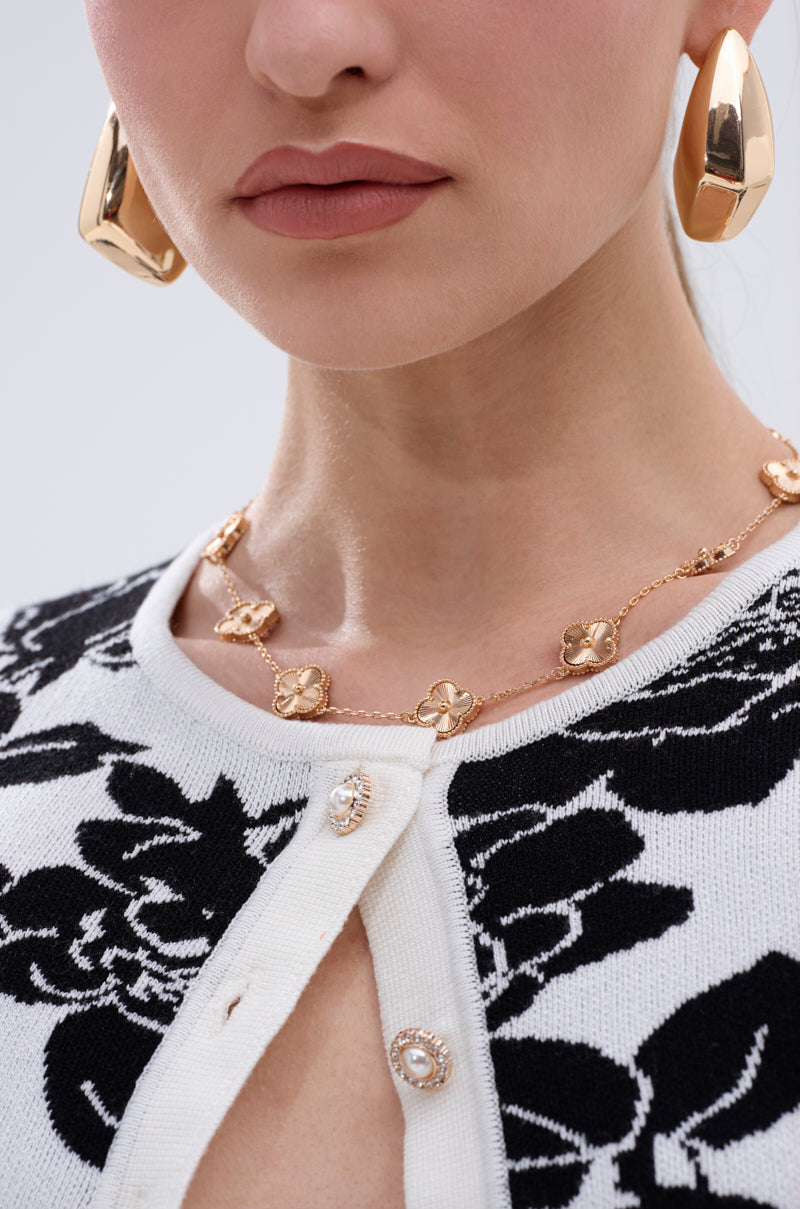 A woman models the QUEZ NECKLACE—a gold-tone piece with floral charms—paired with large gold earrings and a black-and-white patterned cardigan with pearl buttons, shown from the shoulders up for a detailed look at her jewelry and outfit. 3