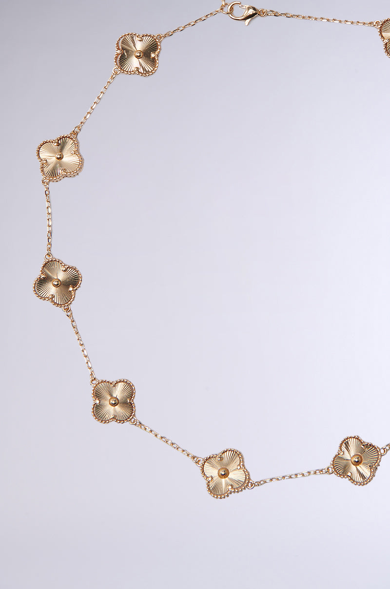 The QUEZ NECKLACE is a gold-tone piece with evenly spaced floral charms on delicate chain links, finished with an adjustable lobster clasp, displayed on a light gray background. 4