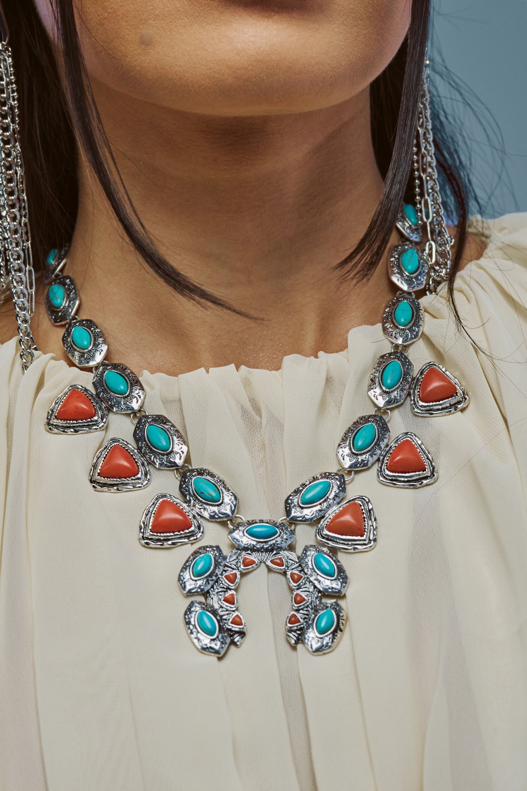 A close-up of a woman wearing a cream blouse and the WAY OUT WEST TURQUOISE NECKLACE, featuring a large silver horseshoe pendant adorned with turquoise and coral-colored stones in an intricate design—making it a striking piece of western jewelry.