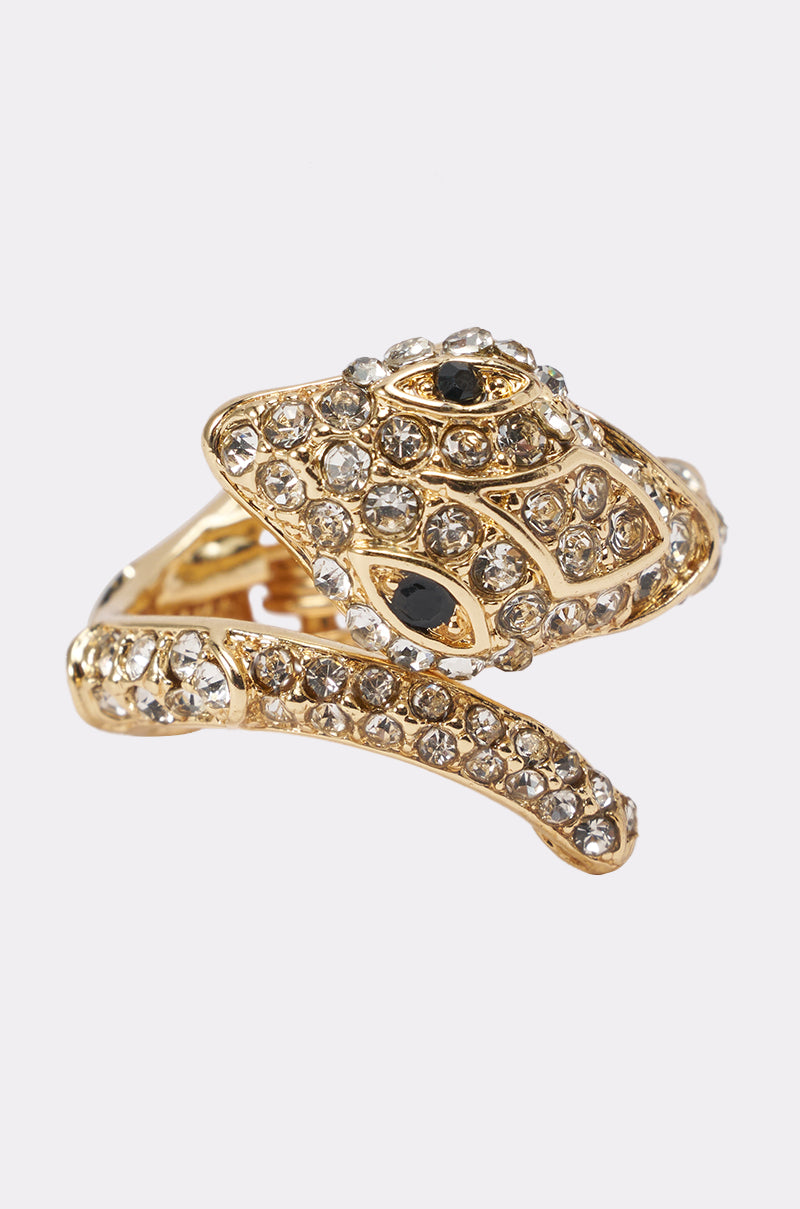 The WRAPPED AROUND YOUR FINGER SNAKE RING features a captivating gold snake shape, coiled in a spiral and adorned with sparkling clear rhinestones and striking black gemstone eyes. Set against a plain white background, this statement jewelry piece perfectly embodies the allure of a bold rhinestone snake ring.
