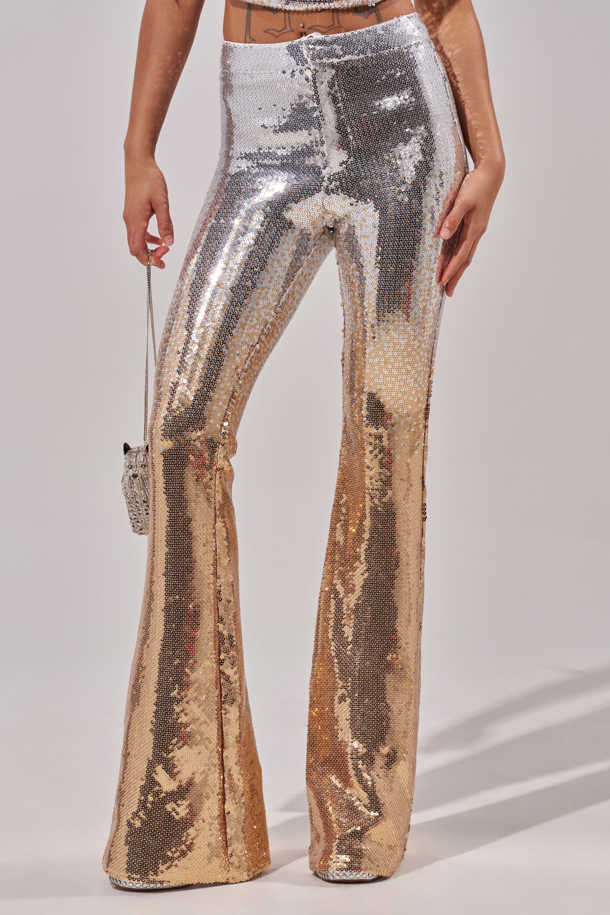 A person wearing GALAXY OMBRE SEQUIN FLARE PANT, which transitions from silver to gold, holds a small matching purse. Only their lower torso and legs are shown.