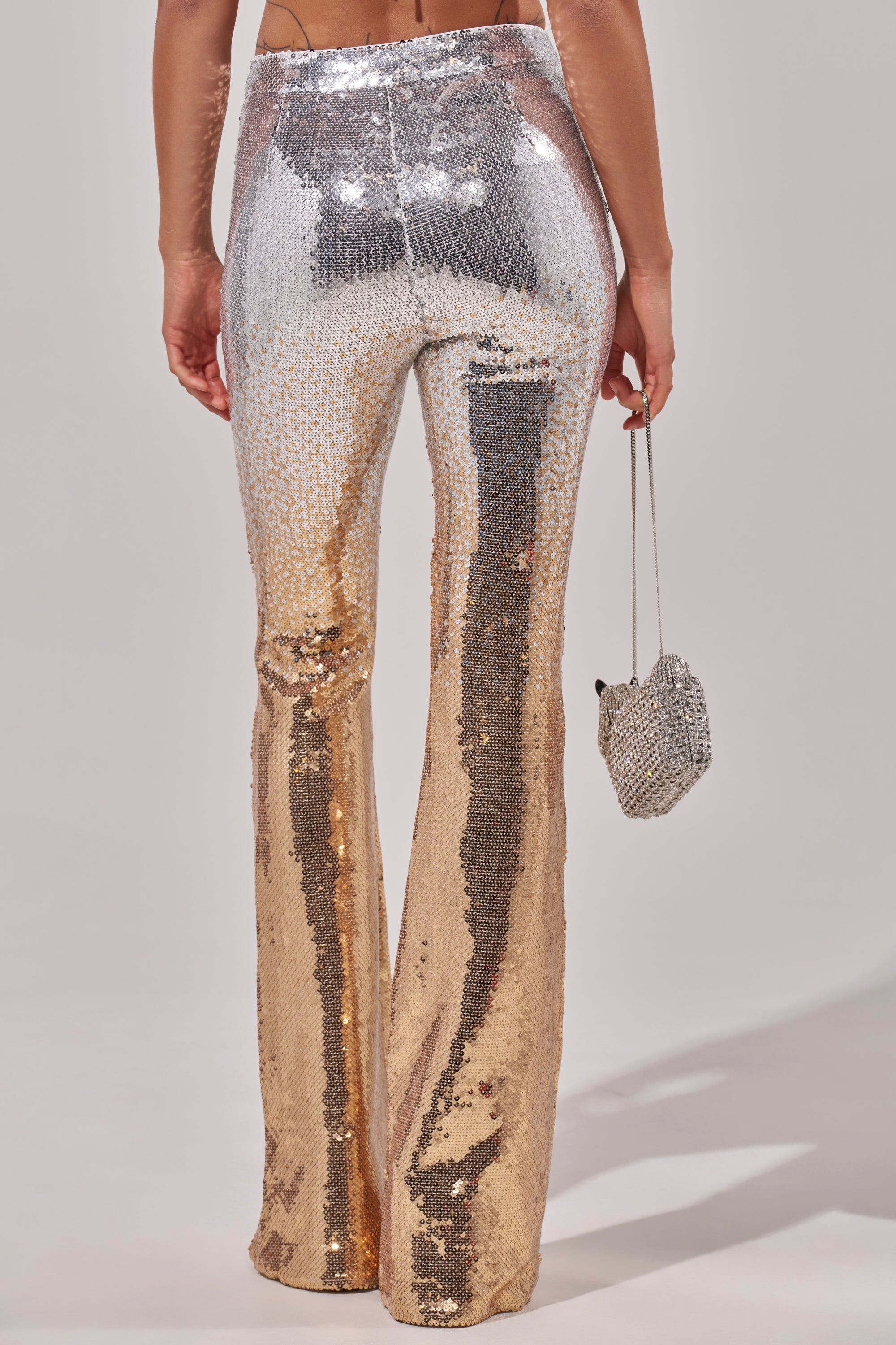 A person wears the GALAXY OMBRE SEQUIN FLARE PANT, featuring a high-waisted fit and silver-to-gold gradient sequins, holding a sparkly silver bag, photographed from behind against a plain background.