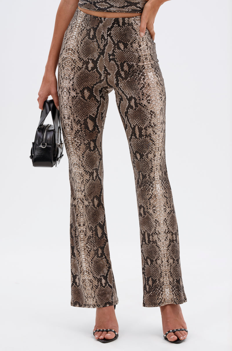 A person wearing LOVE ME HARDER SNAKE FLARED LEGGING in a brown and beige snake print, paired with black heeled sandals and a small black handbag. Only the lower body is visible against a white background.