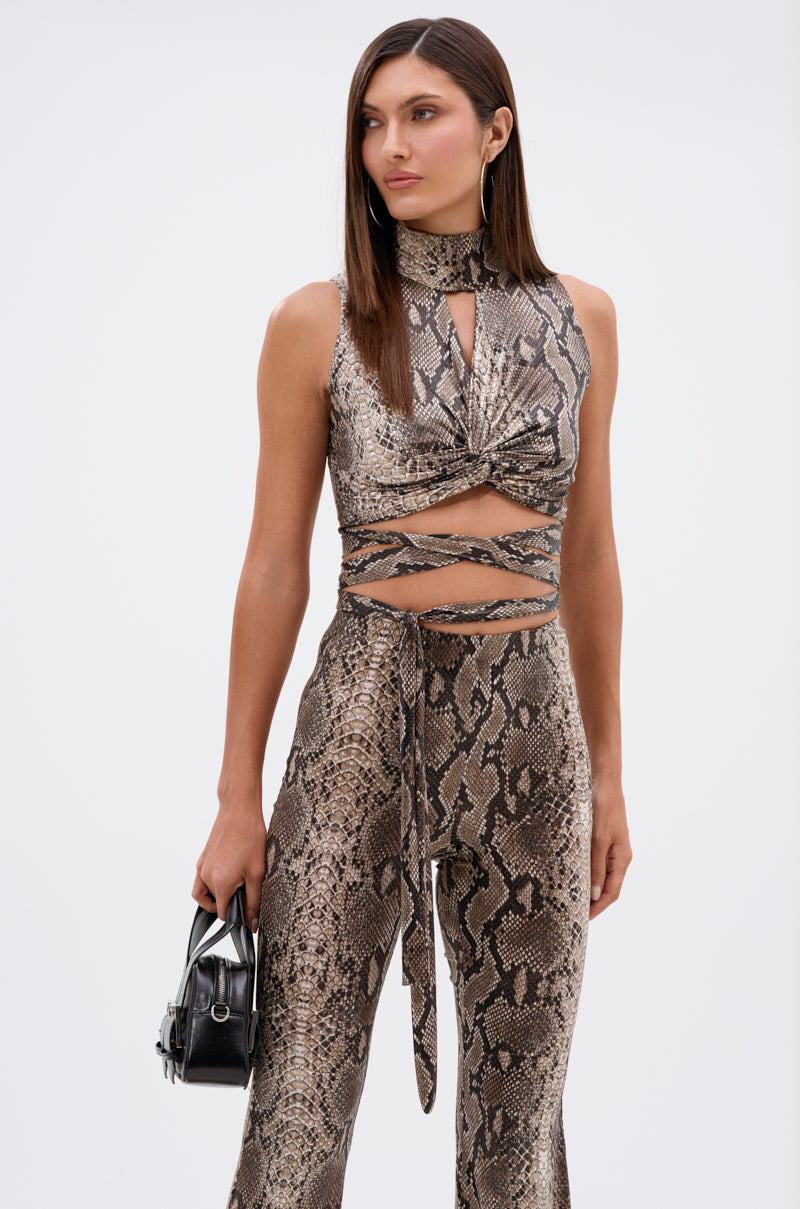 A woman in the LOVE ME HARDER MOCKNECK SNAKE BLOUSE and matching flared pants holds a small black handbag. She has straight, cropped brown hair and stands against a plain white background. 1