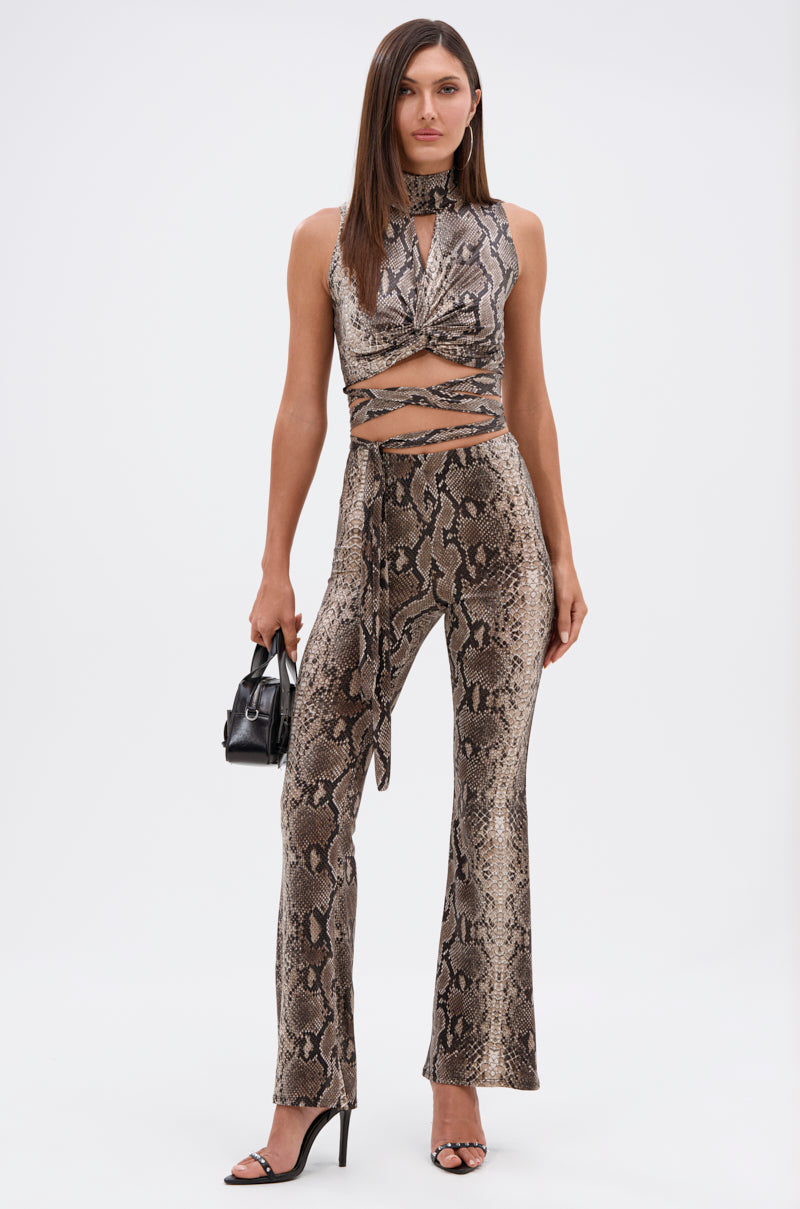 A woman poses in the LOVE ME HARDER MOCKNECK SNAKE BLOUSE, a sleeveless, snakeskin-print cropped top with crisscross wrap details, paired with matching high-waisted pants. She holds a black handbag and wears black heels against a plain white background. 5