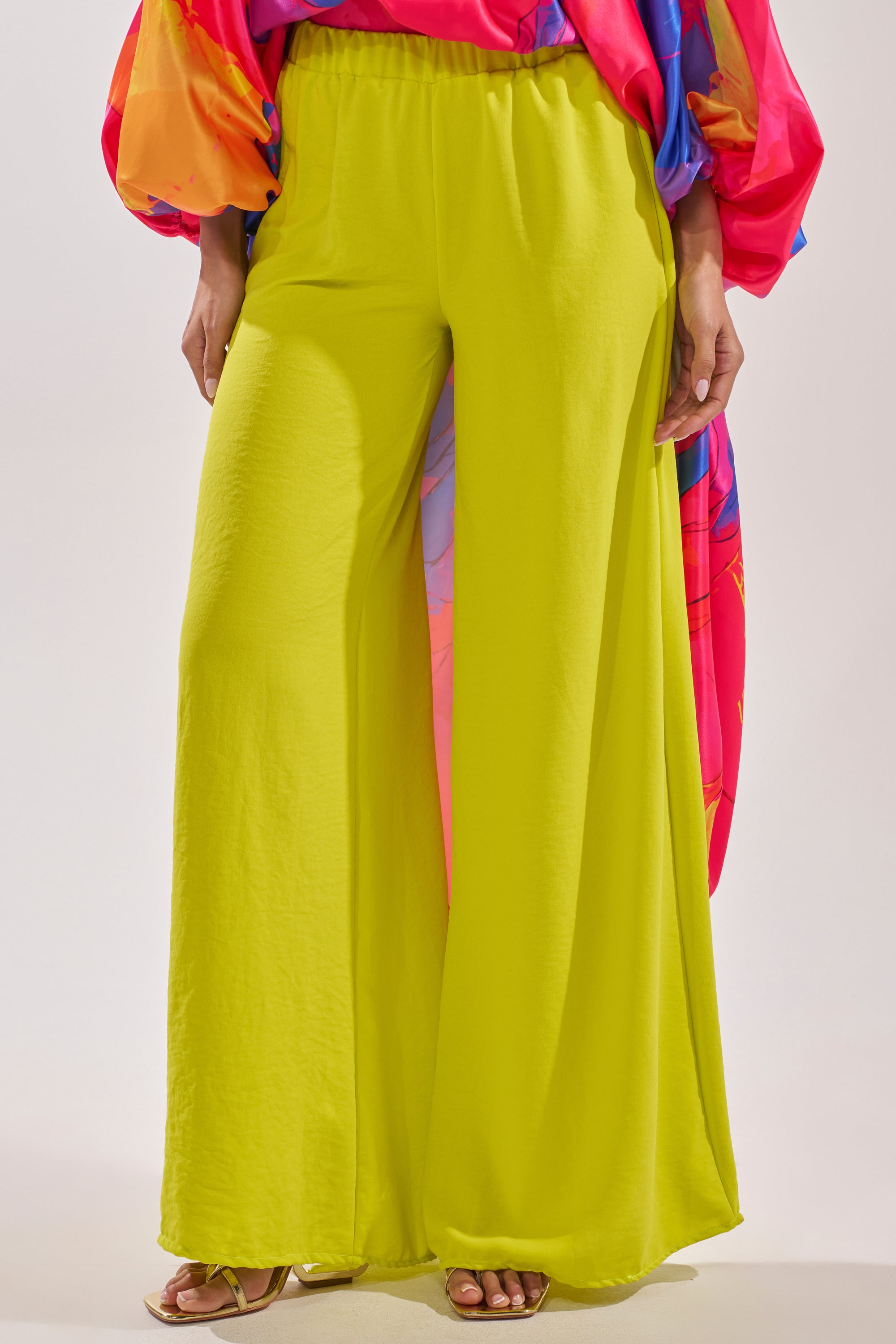A person wearing the BRIGHTEST IN THE CITY PALAZZO PANT, styled with beige open-toe heels and a colorful flowy top featuring pink, blue, and orange patterns. The photo is cropped at the shoulders. 0