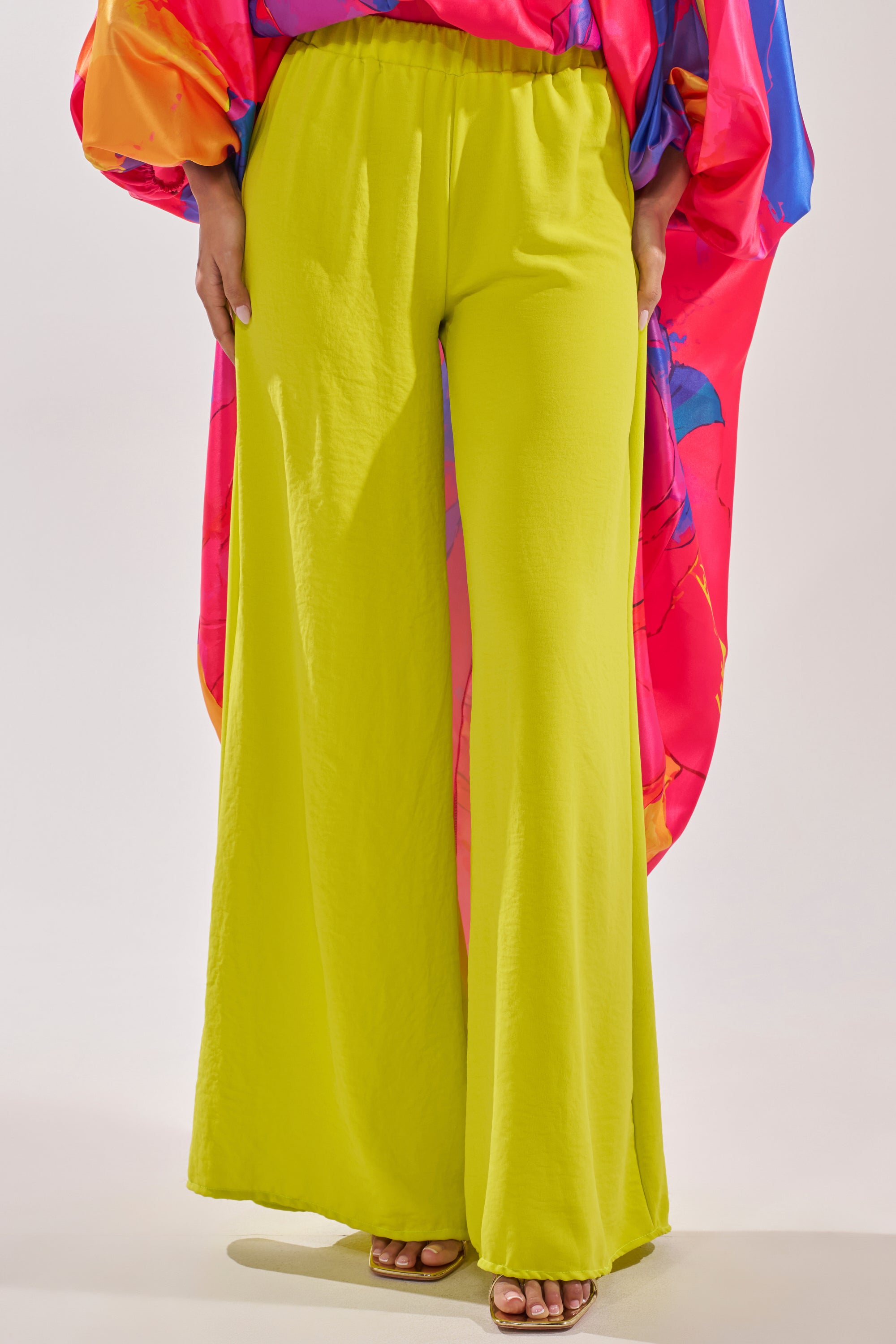 A person models the BRIGHTEST IN THE CITY PALAZZO PANT in bright yellow, styled with open-toe sandals and a loose pink and blue patterned top. The photo focuses on the playful lower half of the outfit. 1