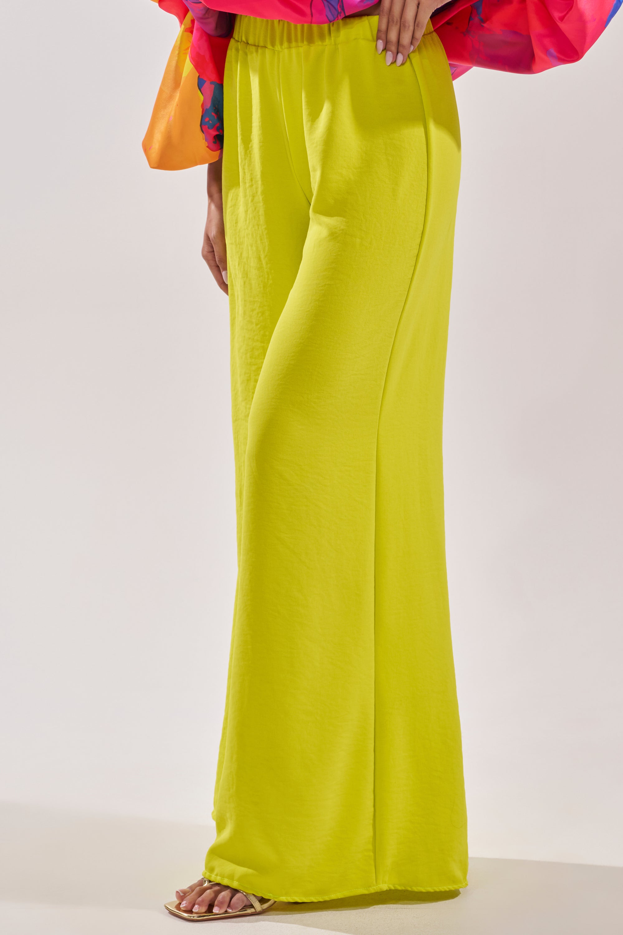 A person showcases the BRIGHTEST IN THE CITY PALAZZO PANT—high-rise and bright yellow—with a colorful long-sleeve top and open-toe sandals, standing against a plain light background. Only the lower body and one hand are visible. 2