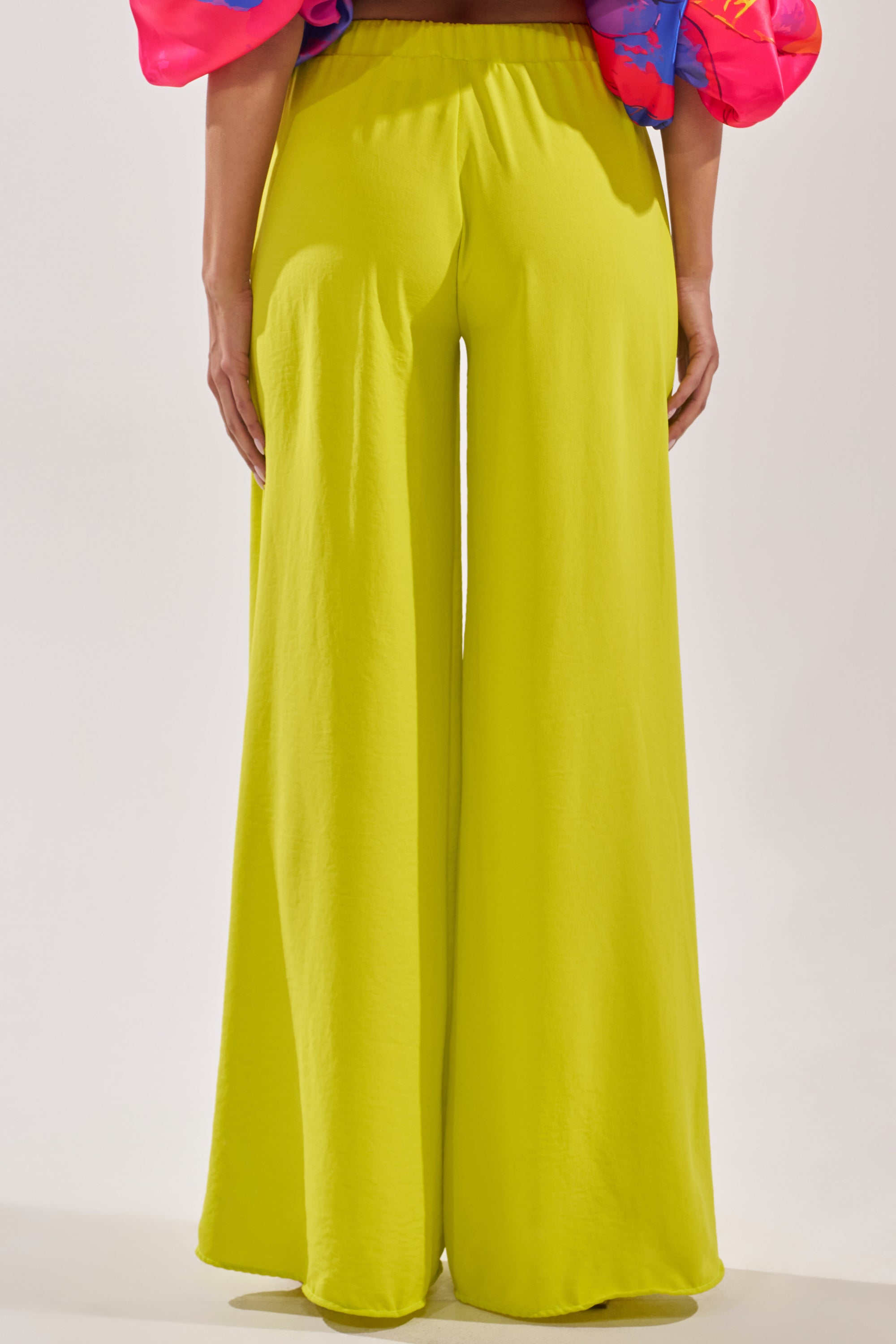 A person stands with their back to the camera, wearing BRIGHTEST IN THE CITY PALAZZO PANT in yellow and a colorful pink puffy-sleeve top. 3