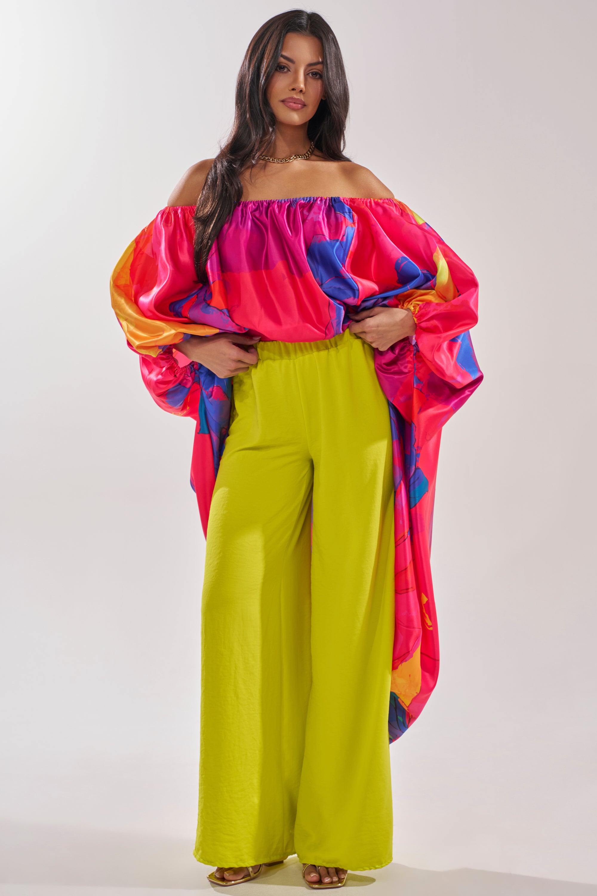 A woman with long dark hair poses confidently against a plain light background, wearing the BRIGHTEST IN THE CITY PALAZZO PANT in lime green and an off-the-shoulder top featuring vibrant pink, blue, and orange patterns. 6
