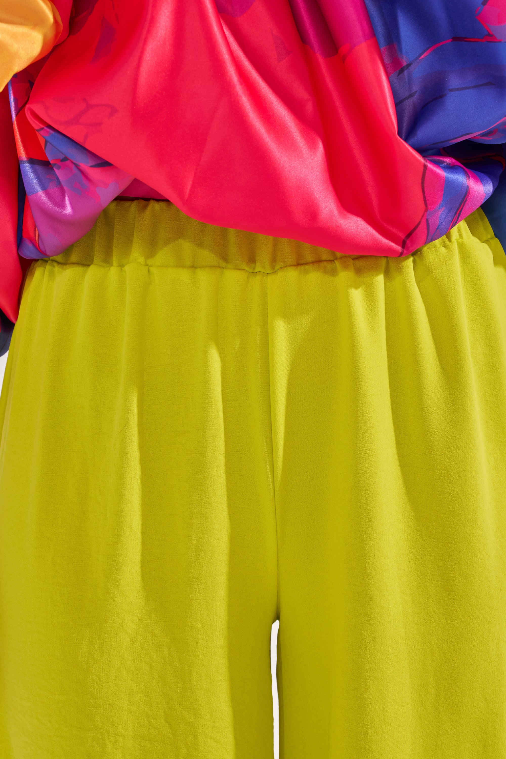 Model wearing BRIGHTEST IN THE CITY PALAZZO PANT in bright yellow, paired with a shiny multicolor top in red, blue, and purple. The image highlights the waist and upper legs; face and full body are not visible. 7