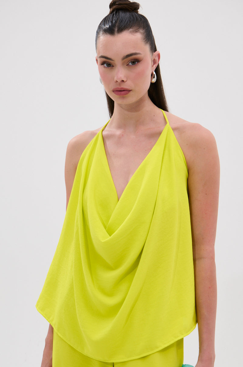 A woman with long dark hair in a high ponytail wears the BRIGHTEST IN THE CITY COWL NECK BLOUSE, a sleeveless neon yellow top with a deep V-neck and open back, made from lightweight fabric, standing against a white background. 0