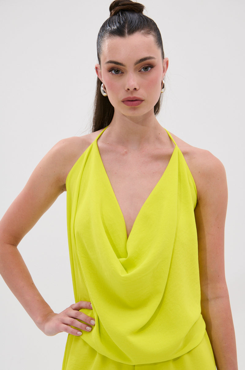 A woman with long dark hair in a high ponytail models the BRIGHTEST IN THE CITY COWL NECK BLOUSE—a sleeveless yellow halter top with a draped neckline and open back—standing against a plain white background with one hand on her hip. 1