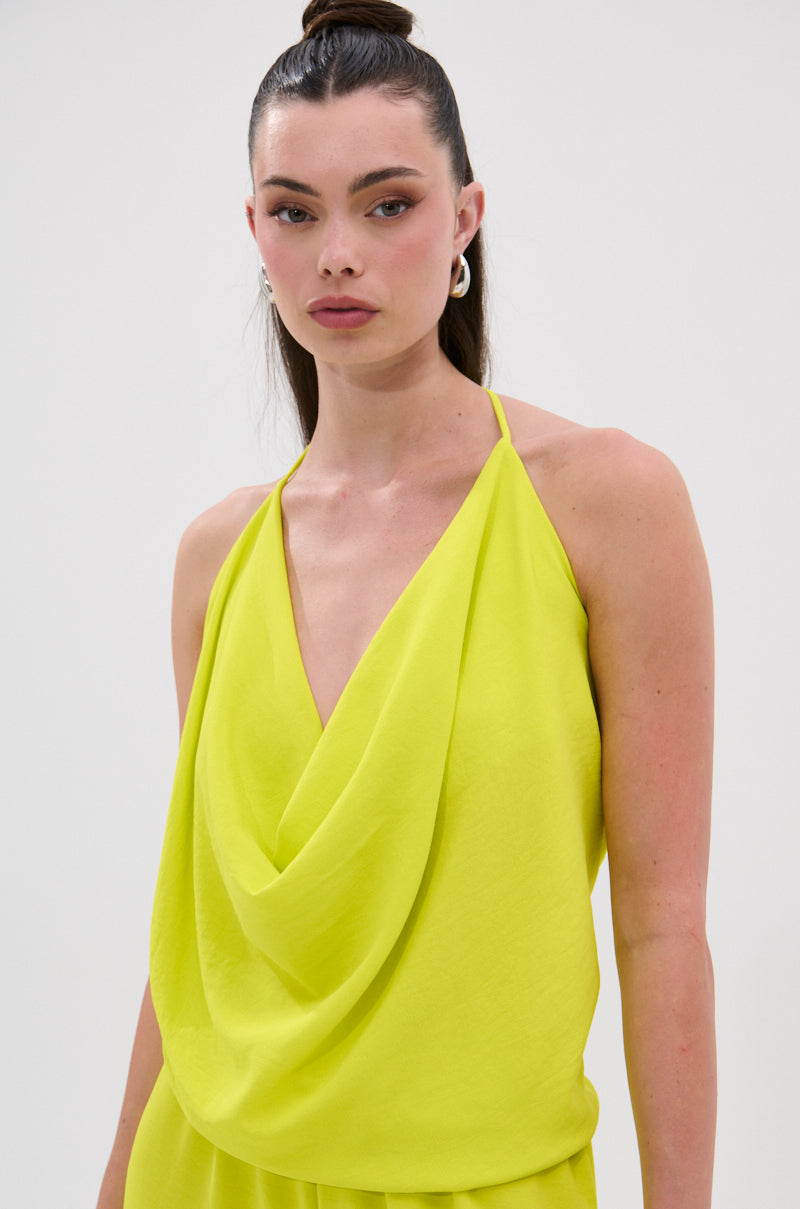 A woman with long brown hair in a high ponytail wears the BRIGHTEST IN THE CITY COWL NECK BLOUSE—a sleeveless, bright yellow draped halter top with an open back—plus silver hoop earrings, standing against a plain light background. 2