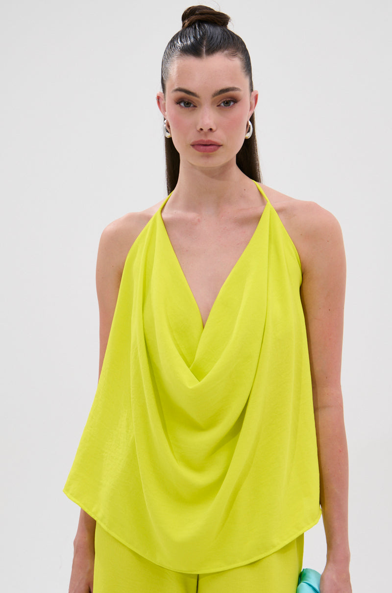 A woman with straight dark hair in a high ponytail wears the BRIGHTEST IN THE CITY COWL NECK BLOUSE, a sleeveless bright yellow draped halter top, standing against a plain light background and looking directly at the camera. 3