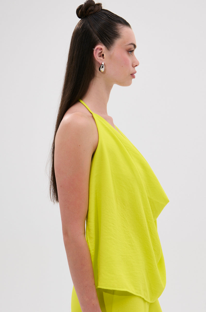 A woman with long straight brown hair in a topknot poses in profile against a white background, wearing the BRIGHTEST IN THE CITY COWL NECK BLOUSE, a sleeveless bright yellow halter top with an open back and matching pants. 4
