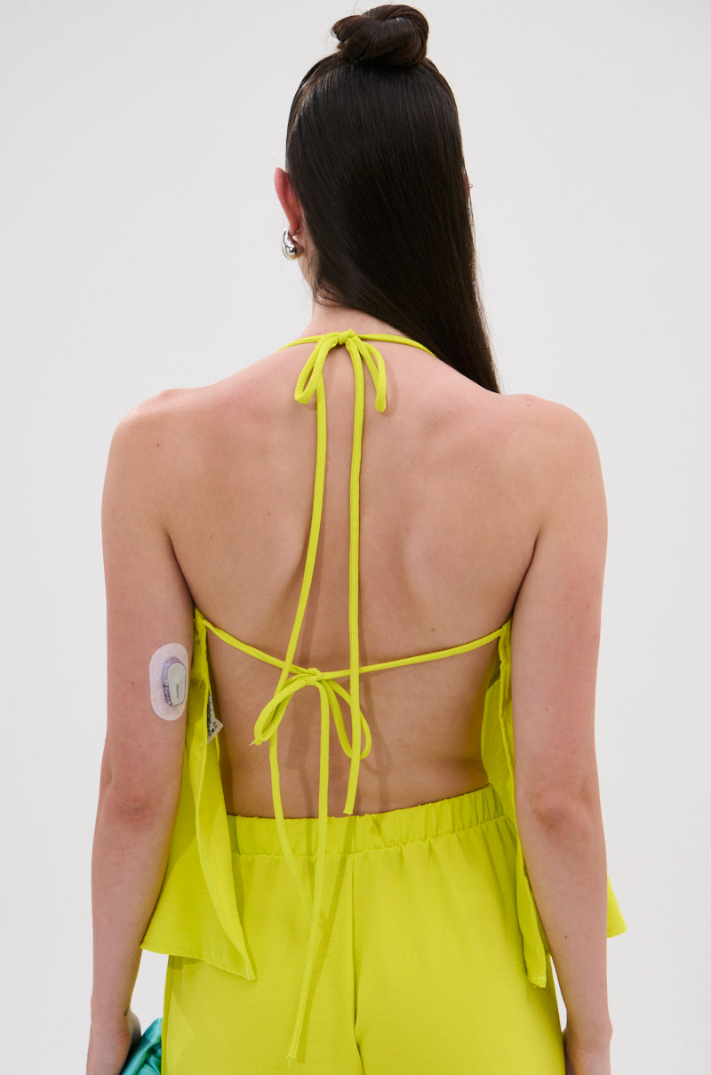 A person with long dark hair wears the BRIGHTEST IN THE CITY COWL NECK BLOUSE in bright yellow, made of lightweight fabric with an open back. A medical device is on their upper left arm, and they hold a turquoise object in their left hand. 5