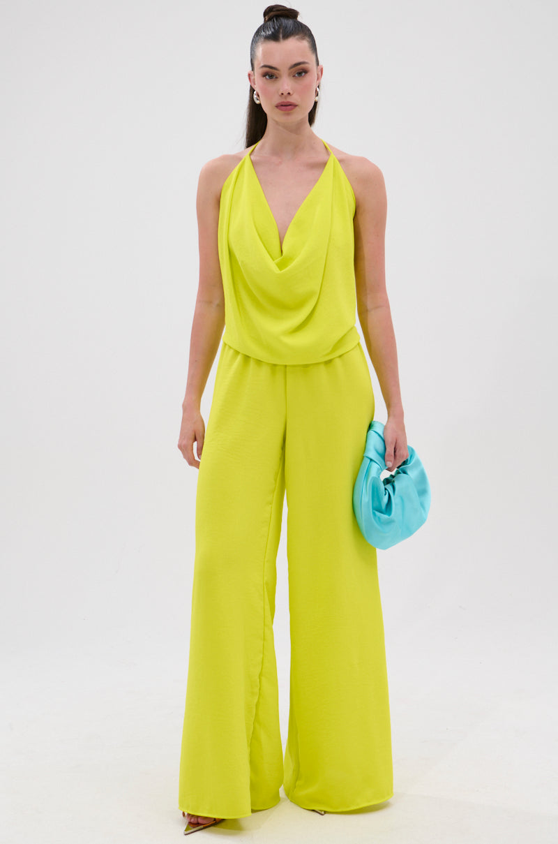 A woman faces forward wearing the BRIGHTEST IN THE CITY COWL NECK BLOUSE, a sleeveless, bright yellow top with a draped neckline. She pairs it with wide-leg pants and a light blue clutch, her hair pulled back against a white background. 6