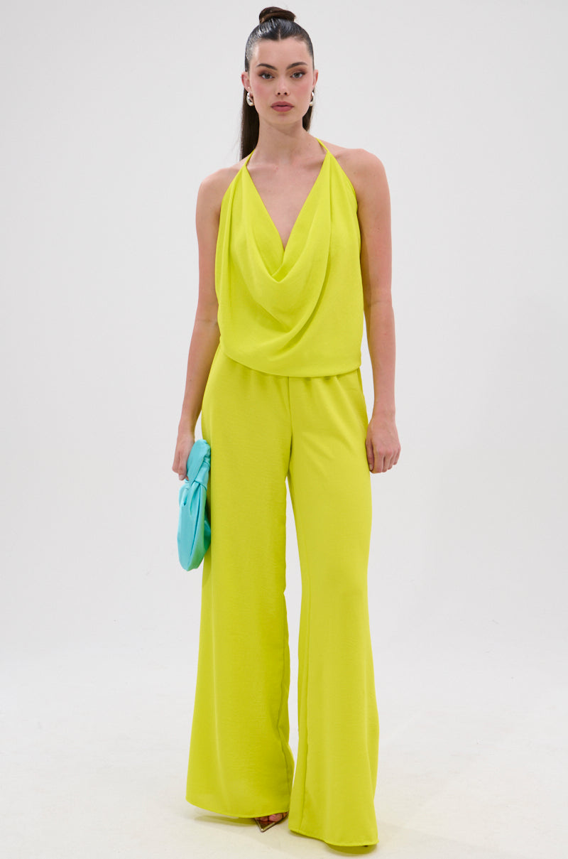 A woman stands against a white background wearing the BRIGHTEST IN THE CITY COWL NECK BLOUSE—a yellow top with a draped cowl neckline and open back. She holds a light blue clutch and styles her hair in a high ponytail. 7