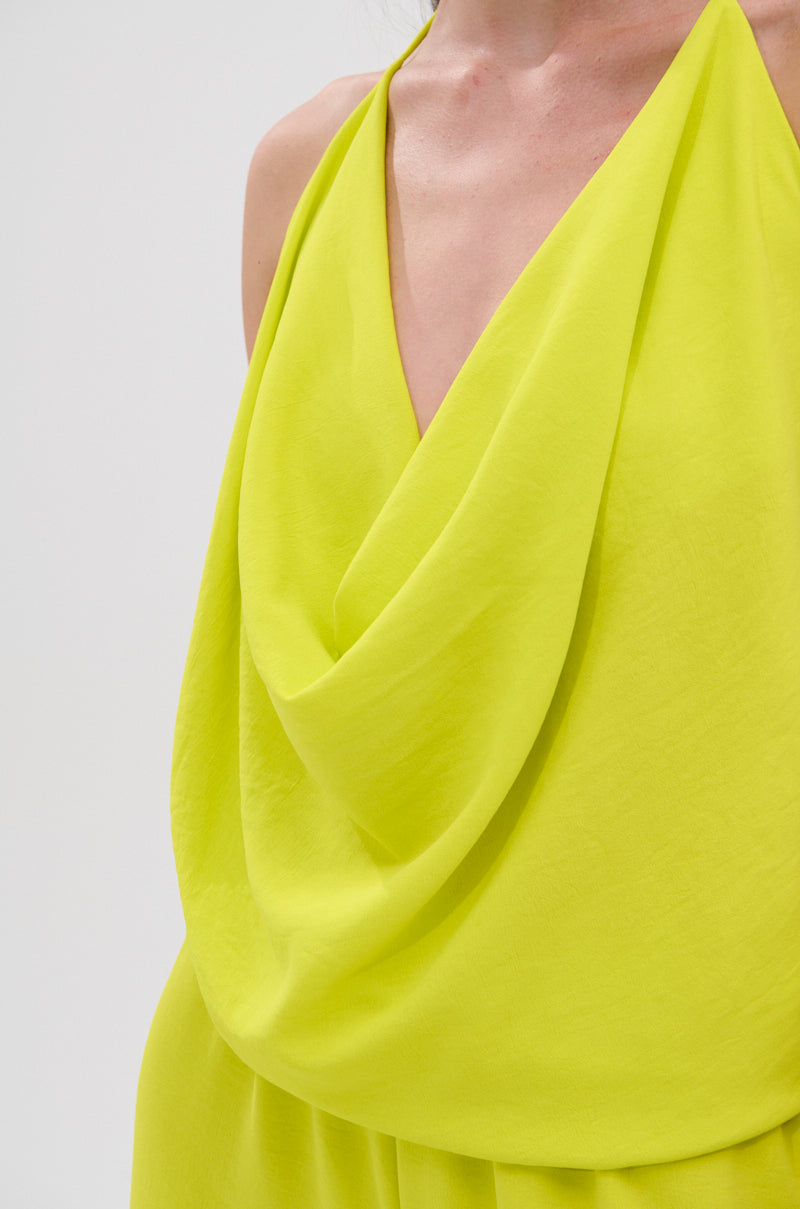 Close-up of a person wearing the BRIGHTEST IN THE CITY COWL NECK BLOUSE, a sleeveless, draped yellow top with a deep cowl neckline. Its lightweight fabric creates soft folds, perfect for standing out against the plain light background. 8
