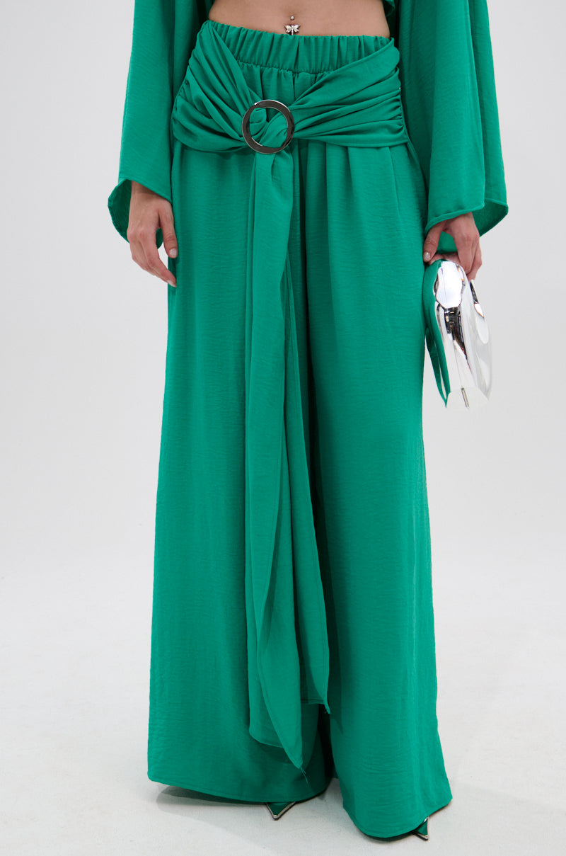 A person wears the CALL ME LATER PALAZZO PANT—loose, high-waisted green pants with a draped, belted detail and circular buckle—while holding a shiny silver clutch against a plain background. 1