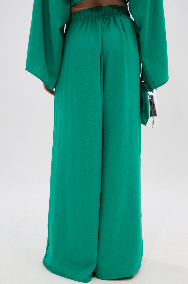 A person models the CALL ME LATER PALAZZO PANT in bright green with a wide-leg, high-rise elastic fit, holding a shiny silver clutch. The waist-down shot is set against a plain background. 3