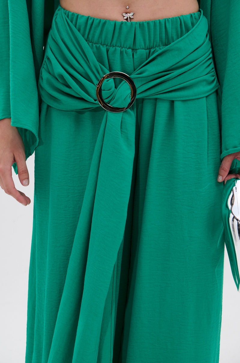 A person wearing the CALL ME LATER PALAZZO PANT in green, featuring a gathered draped waistband with a large metallic buckle and showing a butterfly belly button piercing. Only the lower torso and hands are shown. 5