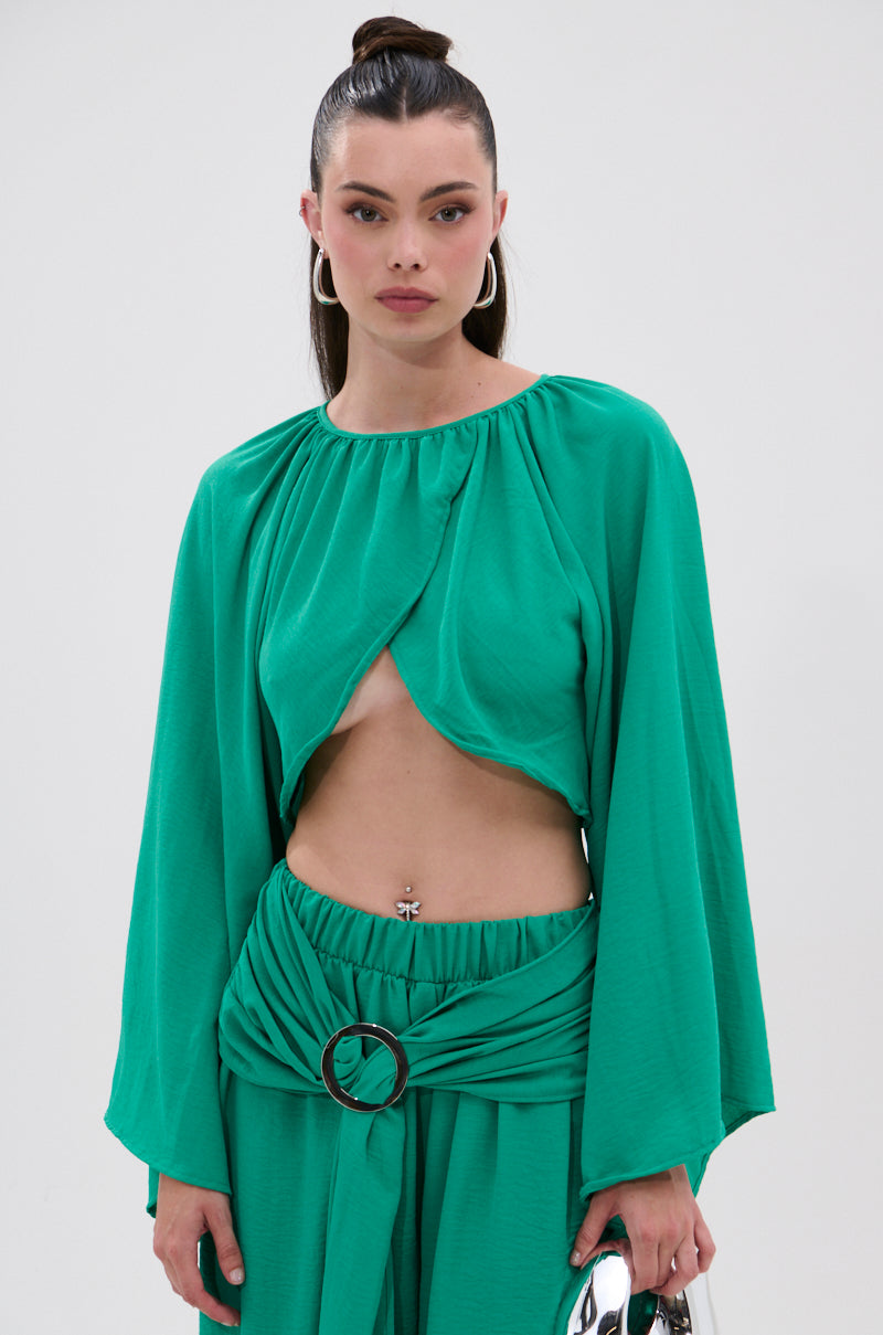 A woman wears the CALL ME LATER CROSSOVER BLOUSE in bright green with long sleeves and a cropped front, paired with matching high-waisted belted pants and large hoop earrings, set against a plain light background.