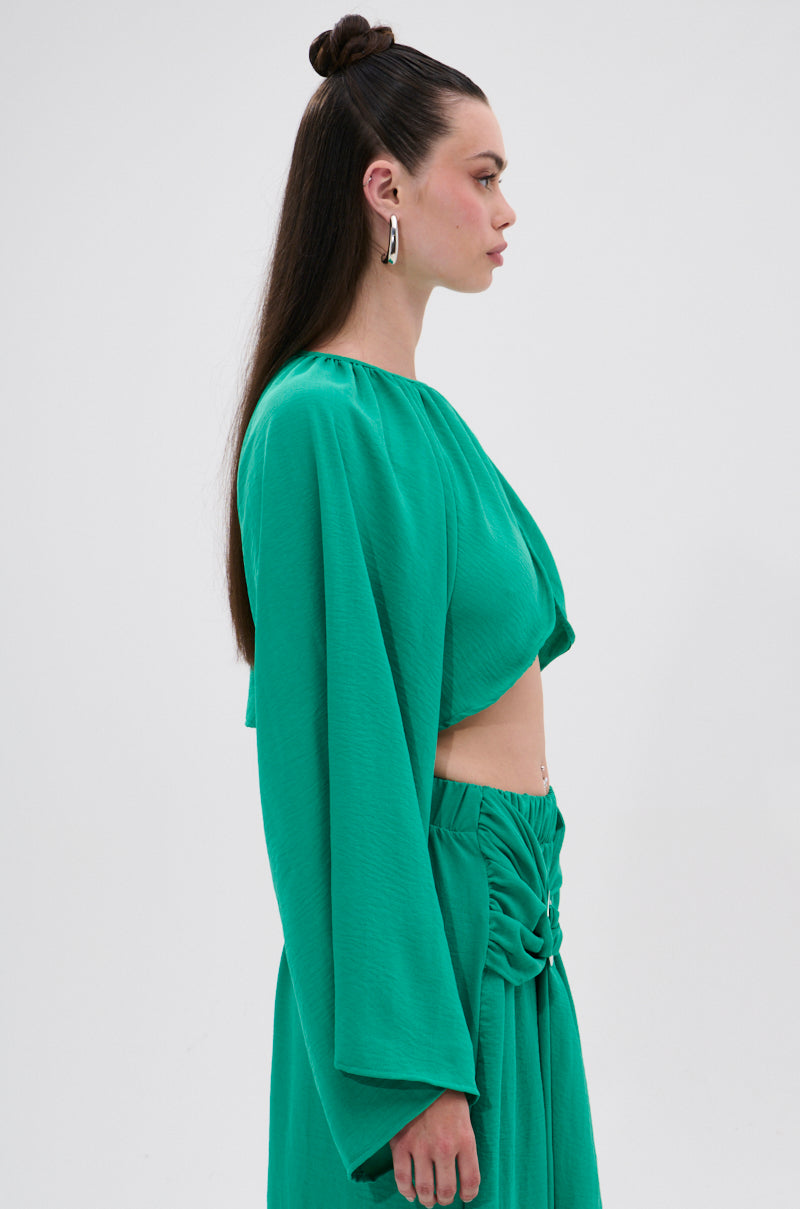 A woman with long, straight dark hair styled half-up wears the vibrant green CALL ME LATER CROSSOVER BLOUSE and a matching high-waisted skirt with gathered details, standing in profile against a plain white background.