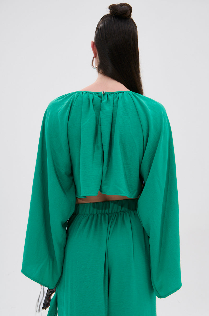 A person with long dark hair is shown from the back wearing the CALL ME LATER CROSSOVER BLOUSE—a vibrant green, lightweight top with wide sleeves, an open back with a small neckline closure, and a chic front crossover.