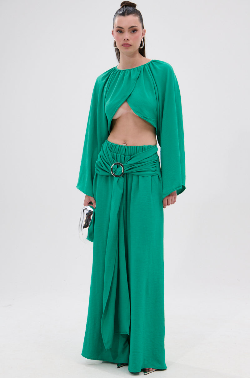 A woman stands against a plain white background wearing the bright green CALL ME LATER CROSSOVER BLOUSE, a two-piece outfit with long sleeves, a midriff cutout, belted wide-leg pants in lightweight fabric, and holding a silver clutch.