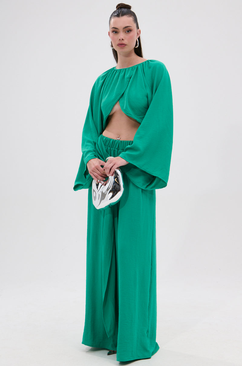 A woman poses against a plain background in the CALL ME LATER CROSSOVER BLOUSE, a vibrant green long-sleeve crop top paired with wide-leg matching pants. She accessorizes with a shiny silver clutch and wears her hair in a high ponytail.